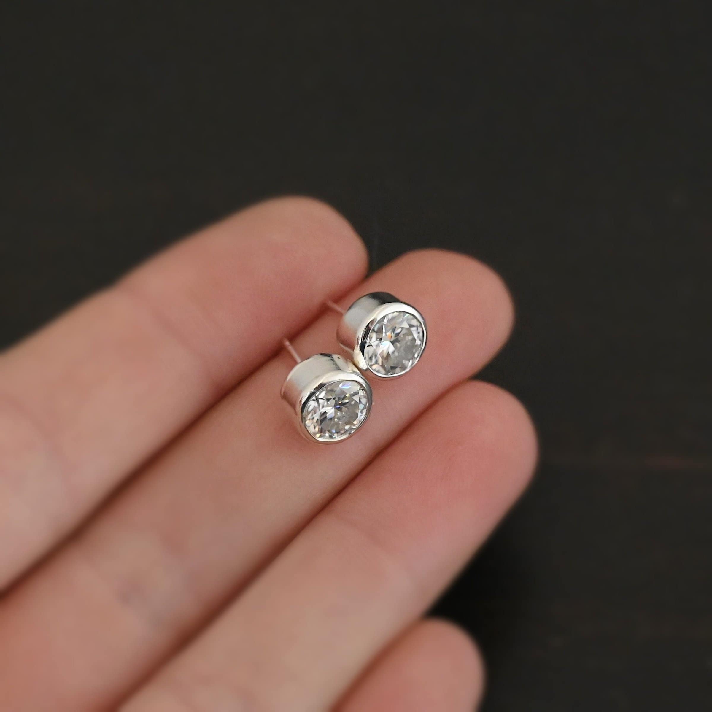 Photo of the "Betina" Earrings. Made out of sterling silver and moissanites. Earrings are sterling silver post earrings with moissanites in the center of them. The settings for the moissanites are thick and the posts are on the back center of the setting. Earrings are held in fingertips of a hand to demonstrate size.