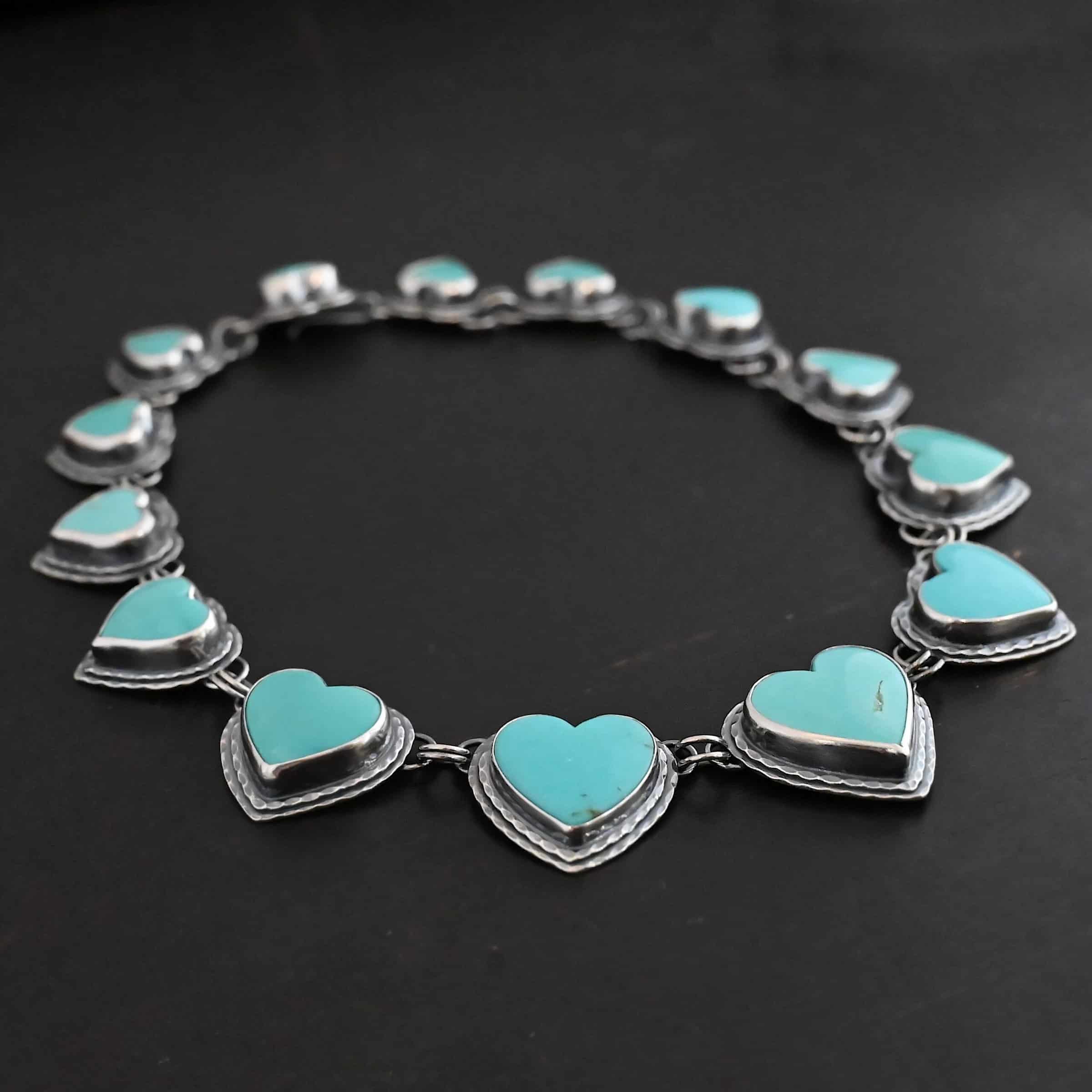Heart & Soul Necklace Close up of the "Heart & Soul" Necklace. Made out of sterling silver and turquoise. There are fourteen turquoise hearts that circle around and create a collar necklace. Each turquoise heart is a light teal color and is set in sterling silver. Necklace is laying against a dark background.