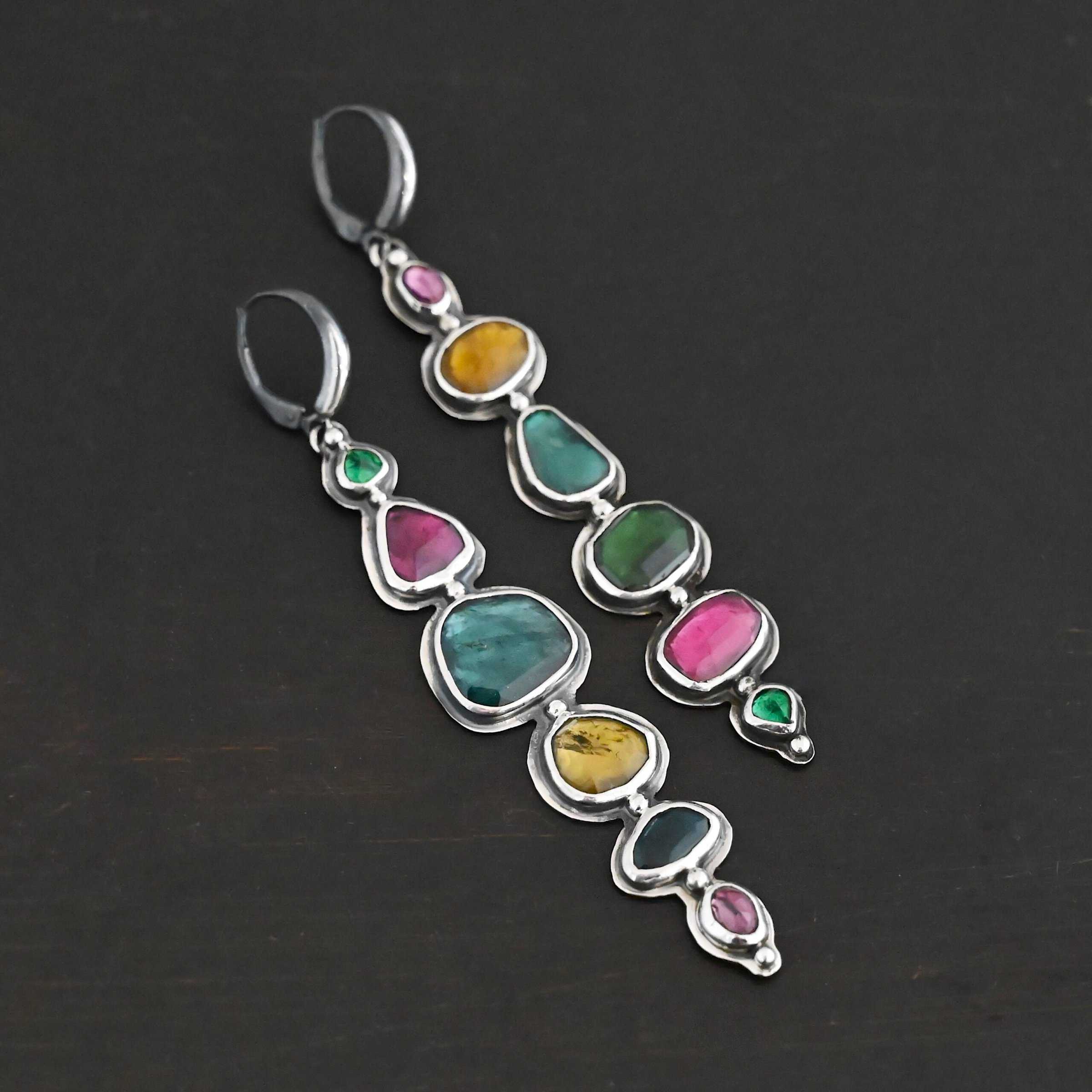 Color Of Love Earrings Close up of the "Color Of Love" Earrings. Made out of sterling silver and sapphires. There are twelve sapphires, six per earring, each a different shape, size, and color. One each earring there are two pink sapphires, two green, one blue, and one yellow. There are seven silver dots on the earring, one on the top of the first stone, five separating each stone afterwards, and one on the bottom of the final stone. Earrings are laying against a dark background.
