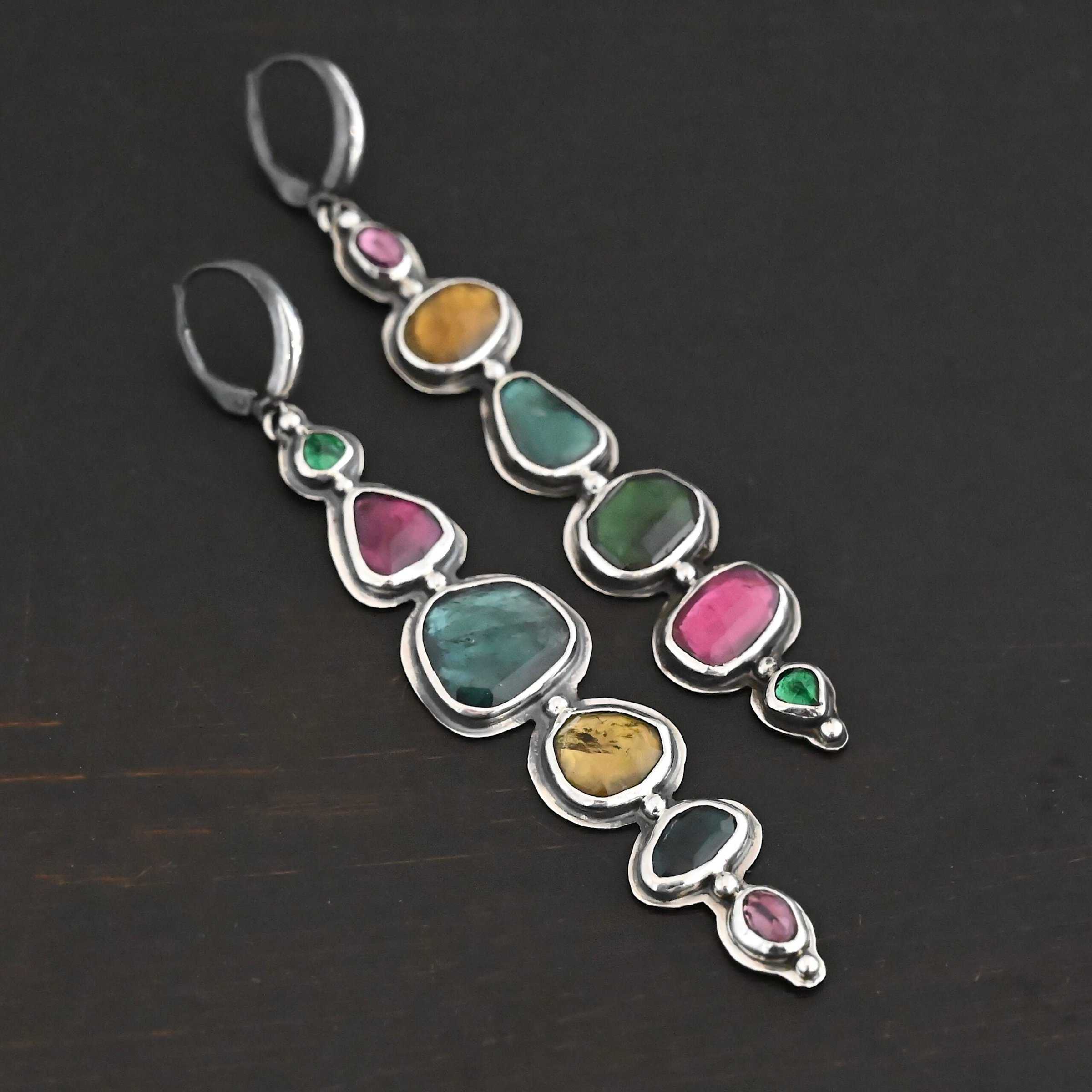 Color Of Love Earrings Close up of the "Color Of Love" Earrings. Made out of sterling silver and sapphires. There are twelve sapphires, six per earring, each a different shape, size, and color. One each earring there are two pink sapphires, two green, one blue, and one yellow. There are seven silver dots on the earring, one on the top of the first stone, five separating each stone afterwards, and one on the bottom of the final stone. Earrings are laying against a dark background.