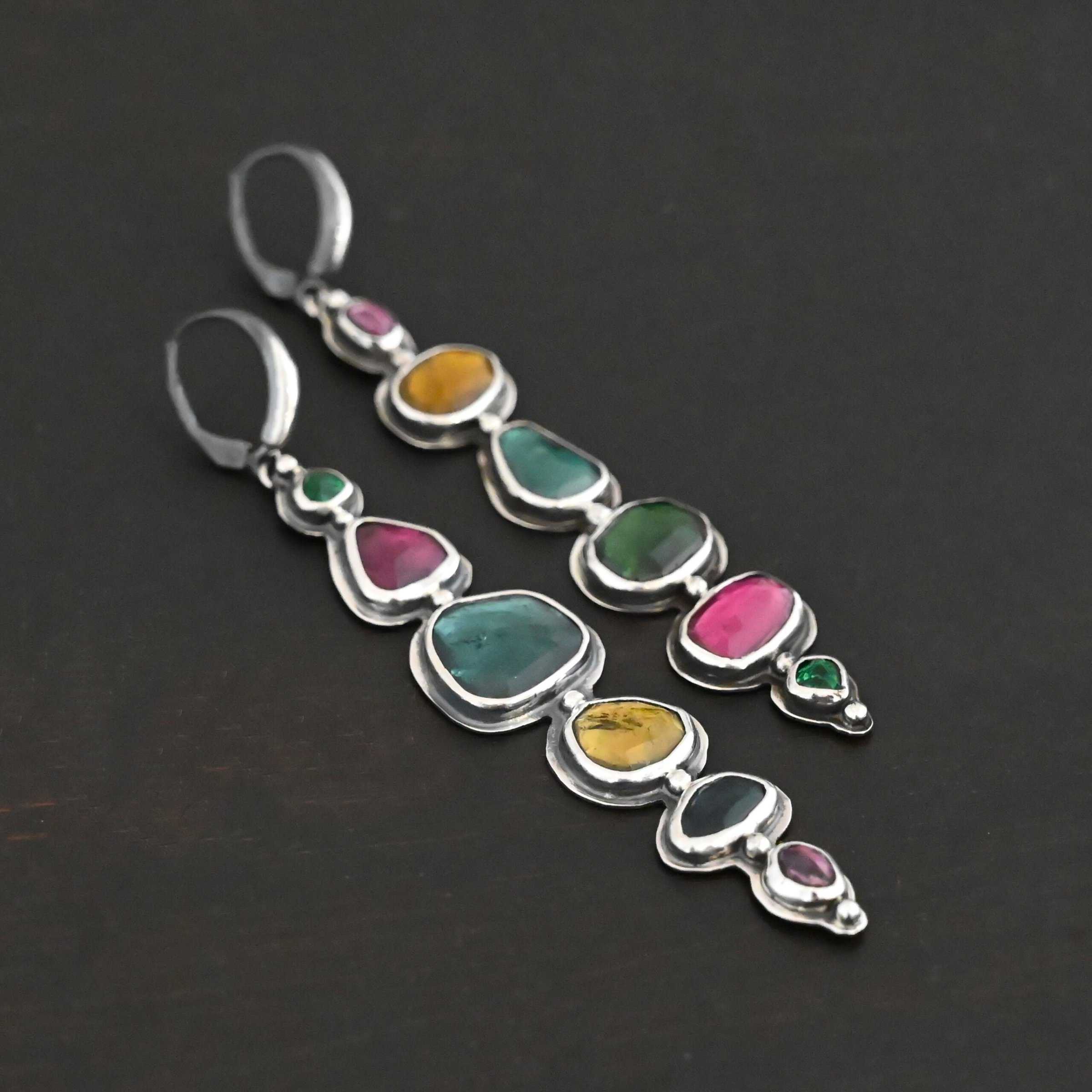 Color Of Love Earrings Close up of the "Color Of Love" Earrings. Made out of sterling silver and sapphires. There are twelve sapphires, six per earring, each a different shape, size, and color. One each earring there are two pink sapphires, two green, one blue, and one yellow. There are seven silver dots on the earring, one on the top of the first stone, five separating each stone afterwards, and one on the bottom of the final stone. Earrings are laying against a dark background.