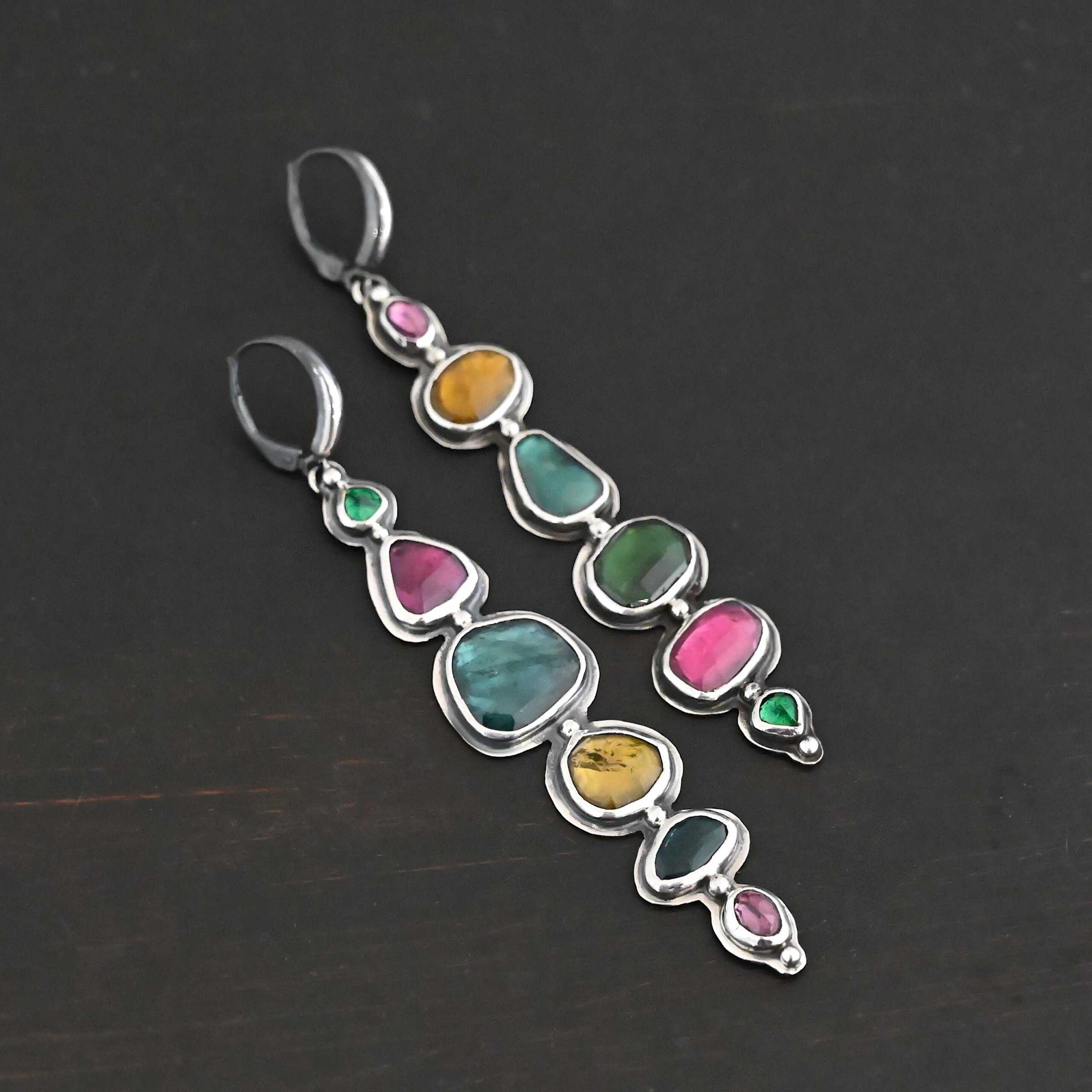 Color Of Love Earrings Close up of the "Color Of Love" Earrings. Made out of sterling silver and sapphires. There are twelve sapphires, six per earring, each a different shape, size, and color. One each earring there are two pink sapphires, two green, one blue, and one yellow. There are seven silver dots on the earring, one on the top of the first stone, five separating each stone afterwards, and one on the bottom of the final stone. Earrings are laying against a dark background.