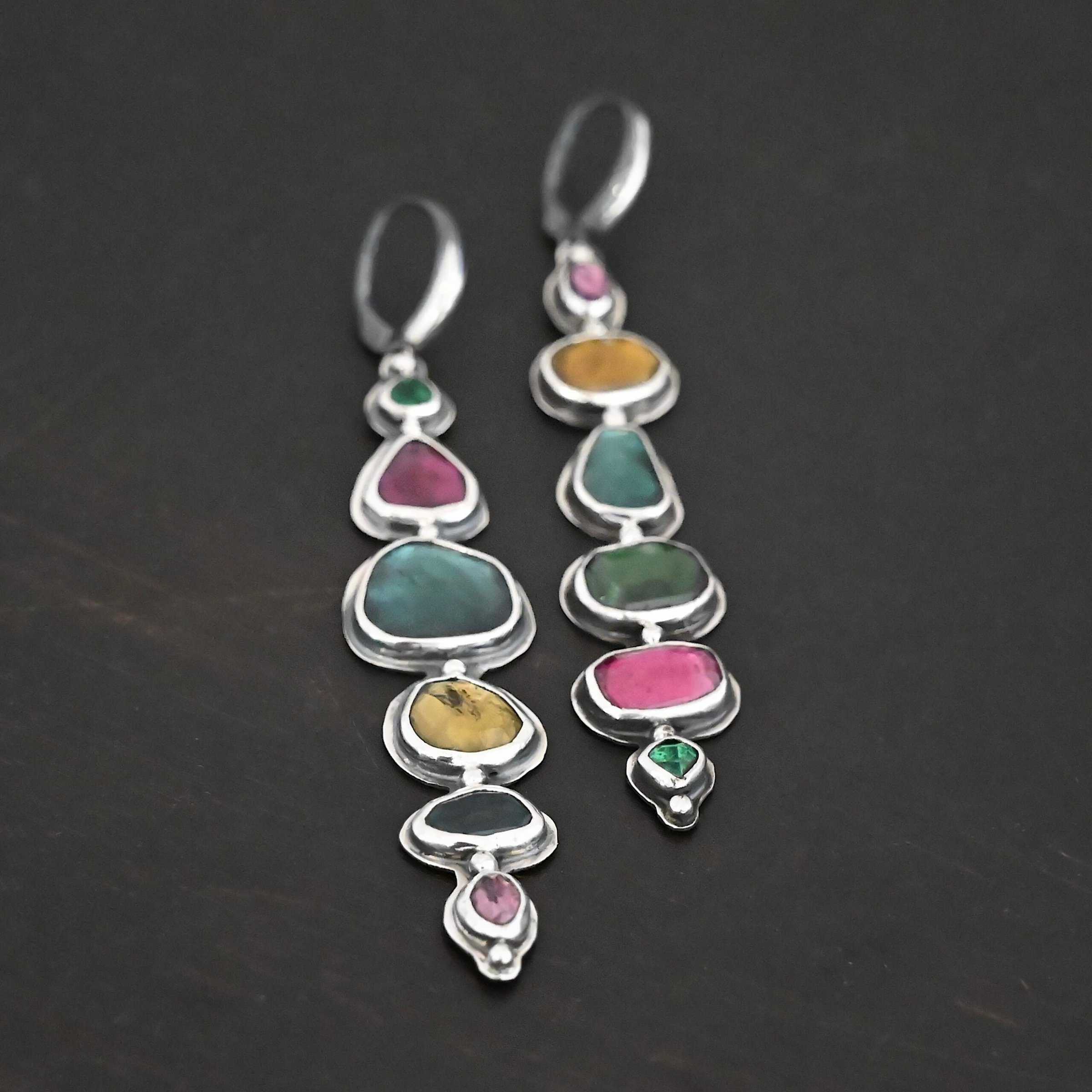Color Of Love Earrings Close up of the "Color Of Love" Earrings. Made out of sterling silver and sapphires. There are twelve sapphires, six per earring, each a different shape, size, and color. One each earring there are two pink sapphires, two green, one blue, and one yellow. There are seven silver dots on the earring, one on the top of the first stone, five separating each stone afterwards, and one on the bottom of the final stone. Earrings are laying against a dark background.