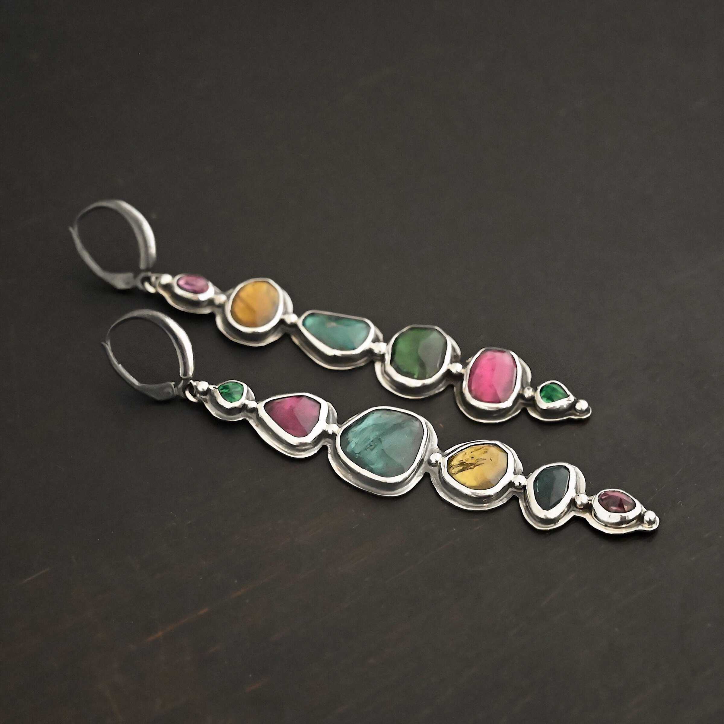 Color Of Love Earrings Close up of the "Color Of Love" Earrings. Made out of sterling silver and sapphires. There are twelve sapphires, six per earring, each a different shape, size, and color. One each earring there are two pink sapphires, two green, one blue, and one yellow. There are seven silver dots on the earring, one on the top of the first stone, five separating each stone afterwards, and one on the bottom of the final stone. Earrings are laying against a dark background.