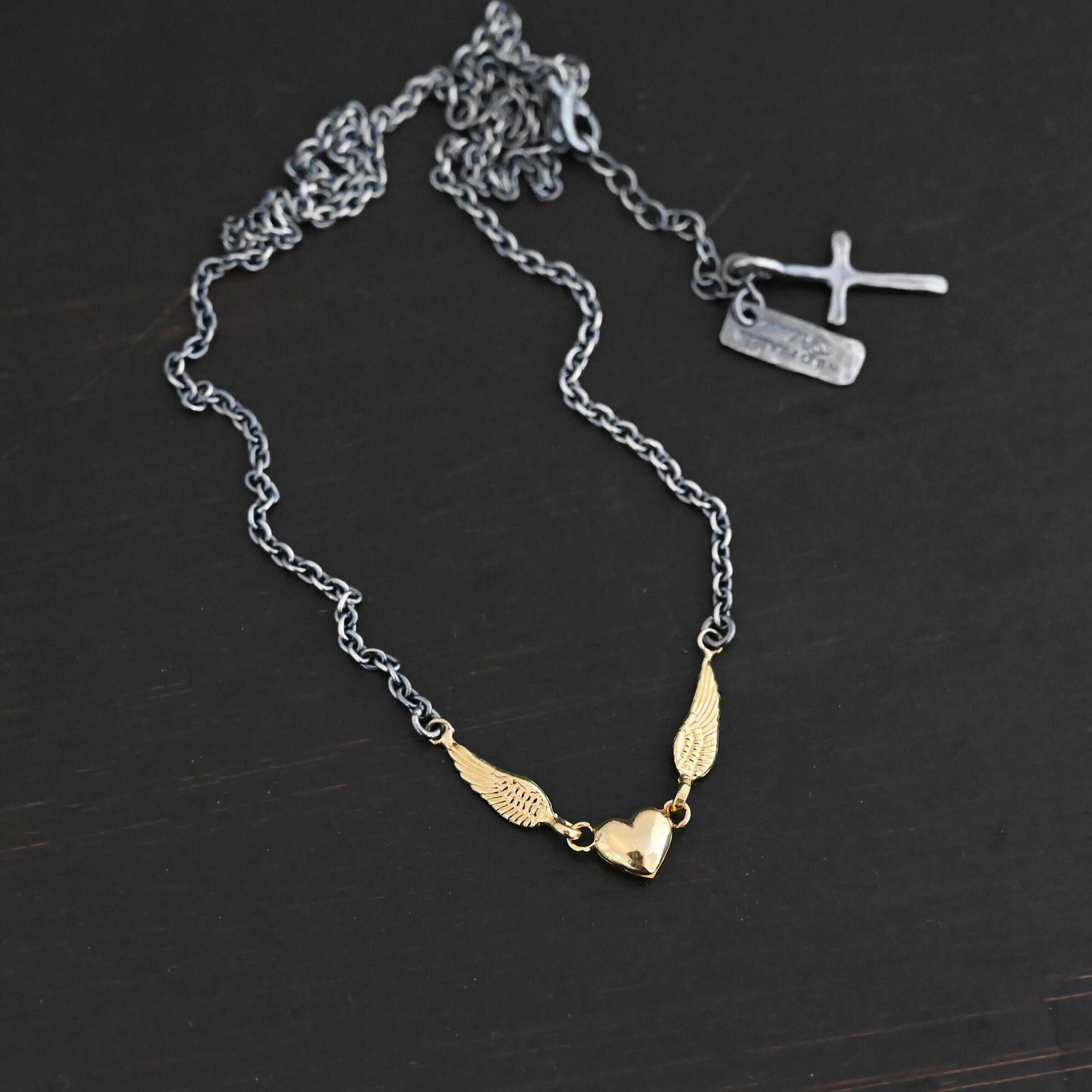 A Little Courage Necklace - Gold Close up of "A Little Courage" Necklace. Made out of sterling silver and 18k gold. There is a small heart in the center of the necklace with two small loops on each side of it. Hanging on each loop are small angel wing charms. These angel wing charms have loops on the end of them in which two jump rings are connected to them. Connected to the jump rings are the chain that create the rest of the necklace. The heart and angel wings are made out of 18k gold and the chain of the necklace is made out of sterling silver. Necklace is laying against a dark background.