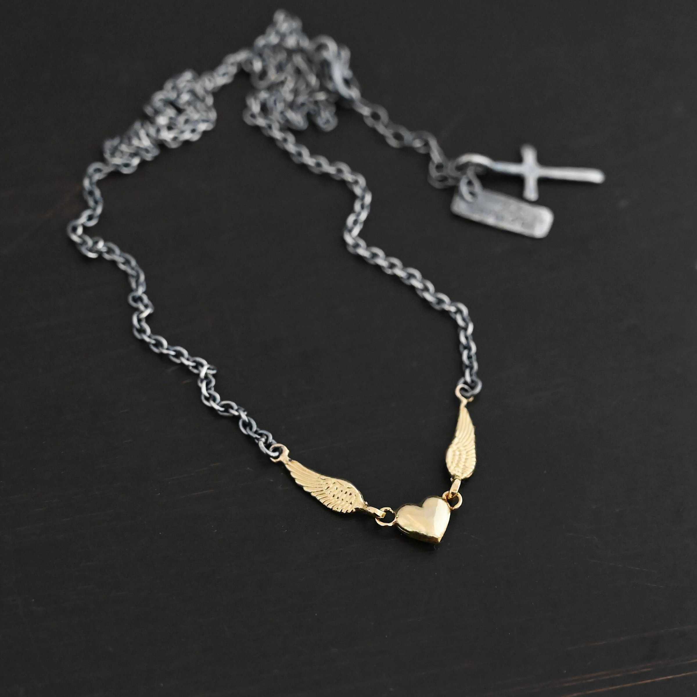 A Little Courage Necklace - Gold Close up of "A Little Courage" Necklace. Made out of sterling silver and 18k gold. There is a small heart in the center of the necklace with two small loops on each side of it. Hanging on each loop are small angel wing charms. These angel wing charms have loops on the end of them in which two jump rings are connected to them. Connected to the jump rings are the chain that create the rest of the necklace. The heart and angel wings are made out of 18k gold and the chain of the necklace is made out of sterling silver. Necklace is laying against a dark background.