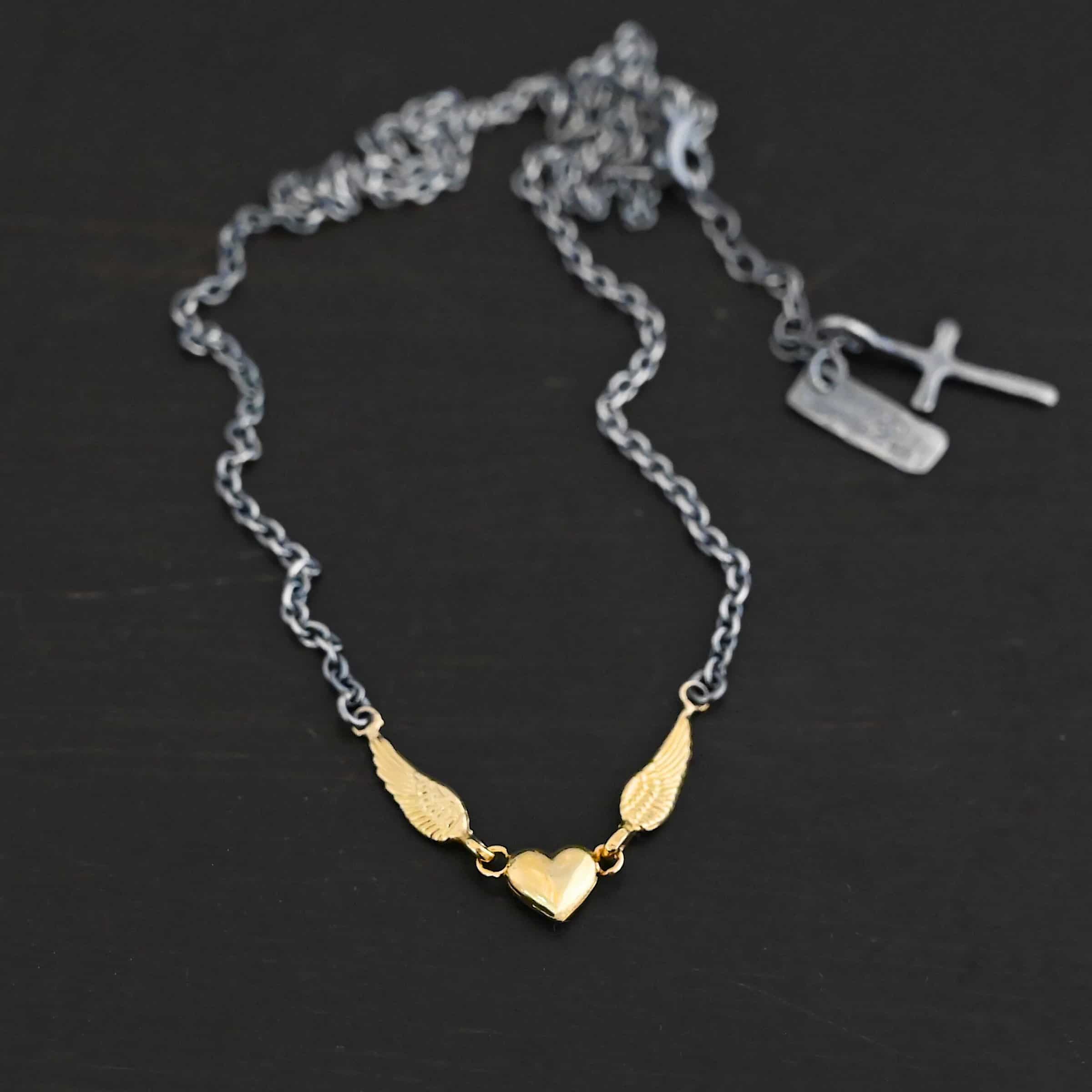A Little Courage Necklace - Gold Close up of "A Little Courage" Necklace. Made out of sterling silver and 18k gold. There is a small heart in the center of the necklace with two small loops on each side of it. Hanging on each loop are small angel wing charms. These angel wing charms have loops on the end of them in which two jump rings are connected to them. Connected to the jump rings are the chain that create the rest of the necklace. The heart and angel wings are made out of 18k gold and the chain of the necklace is made out of sterling silver. Necklace is laying against a dark background.