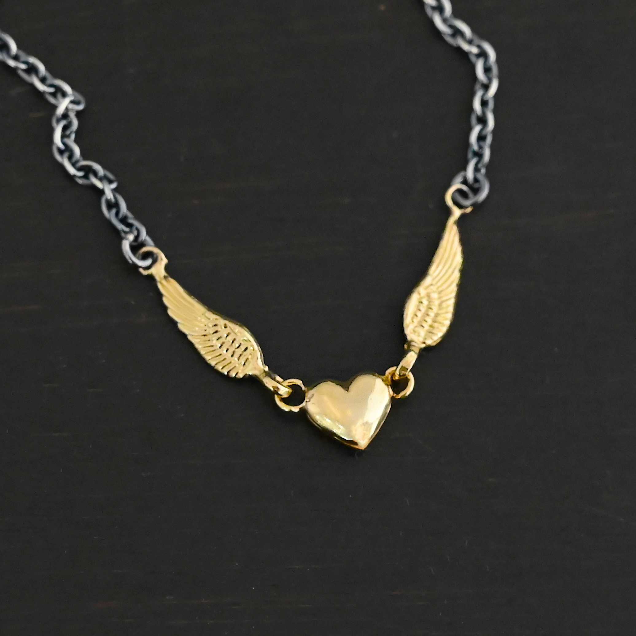 A Little Courage Necklace - Gold Close up of "A Little Courage" Necklace. Made out of sterling silver and 18k gold. There is a small heart in the center of the necklace with two small loops on each side of it. Hanging on each loop are small angel wing charms. These angel wing charms have loops on the end of them in which two jump rings are connected to them. Connected to the jump rings are the chain that create the rest of the necklace. The heart and angel wings are made out of 18k gold and the chain of the necklace is made out of sterling silver. Necklace is laying against a dark background.