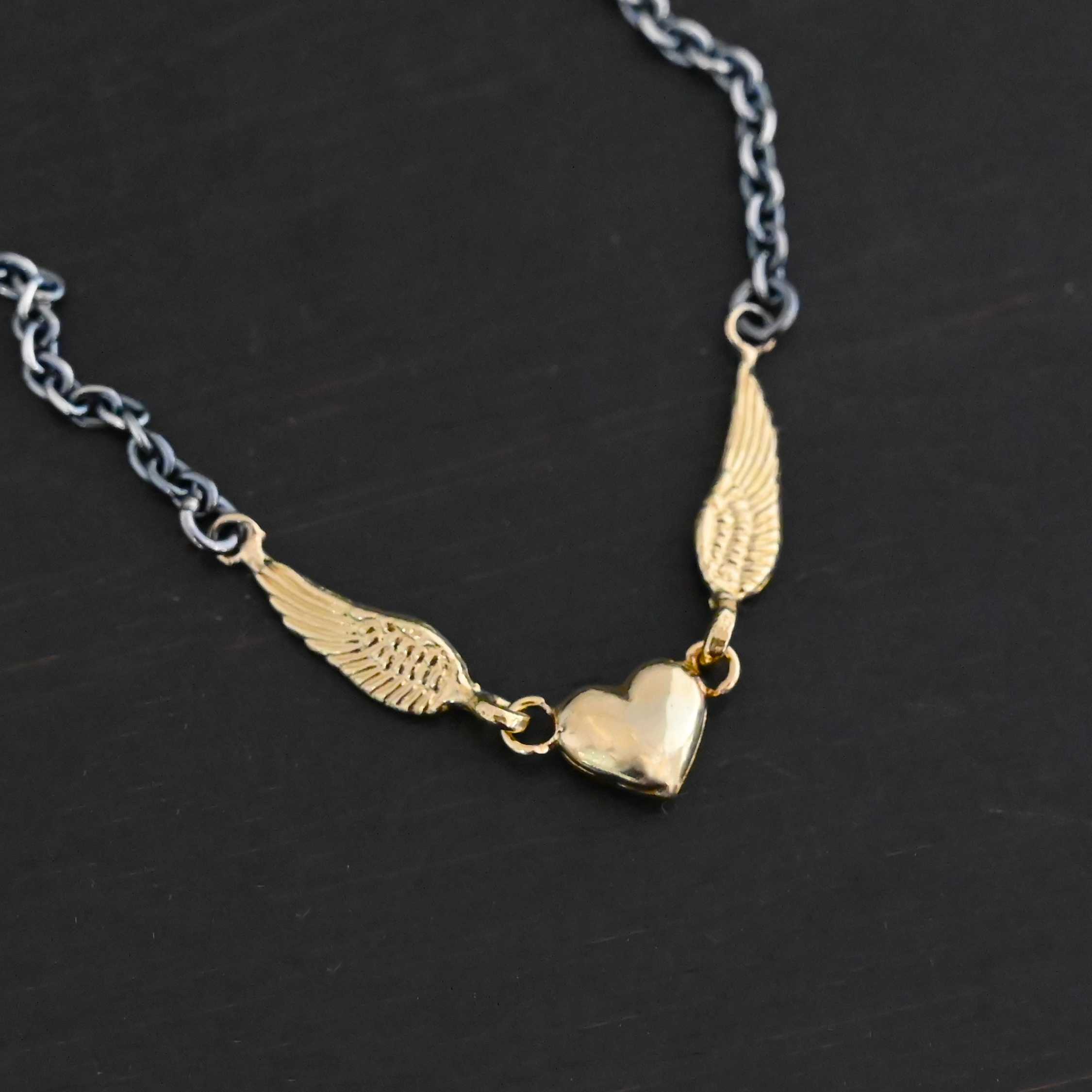 A Little Courage Necklace - Gold Close up of "A Little Courage" Necklace. Made out of sterling silver and 18k gold. There is a small heart in the center of the necklace with two small loops on each side of it. Hanging on each loop are small angel wing charms. These angel wing charms have loops on the end of them in which two jump rings are connected to them. Connected to the jump rings are the chain that create the rest of the necklace. The heart and angel wings are made out of 18k gold and the chain of the necklace is made out of sterling silver. Necklace is laying against a dark background.