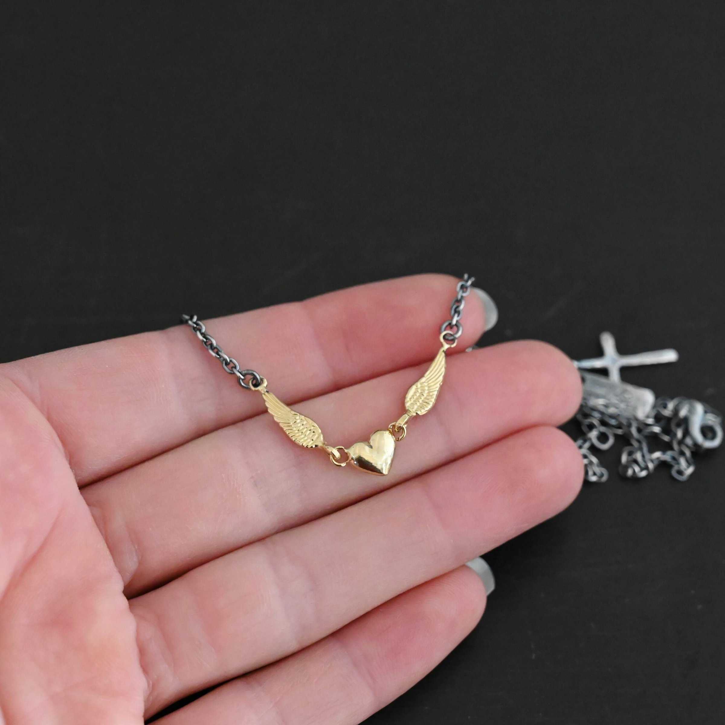 A Little Courage Necklace - Gold Close up of "A Little Courage" Necklace. Made out of sterling silver and 18k gold. There is a small heart in the center of the necklace with two small loops on each side of it. Hanging on each loop are small angel wing charms. These angel wing charms have loops on the end of them in which two jump rings are connected to them. Connected to the jump rings are the chain that create the rest of the necklace. The heart and angel wings are made out of 18k gold and the chain of the necklace is made out of sterling silver. Necklace is laying on the fingertips of a hand to demonstrate size.