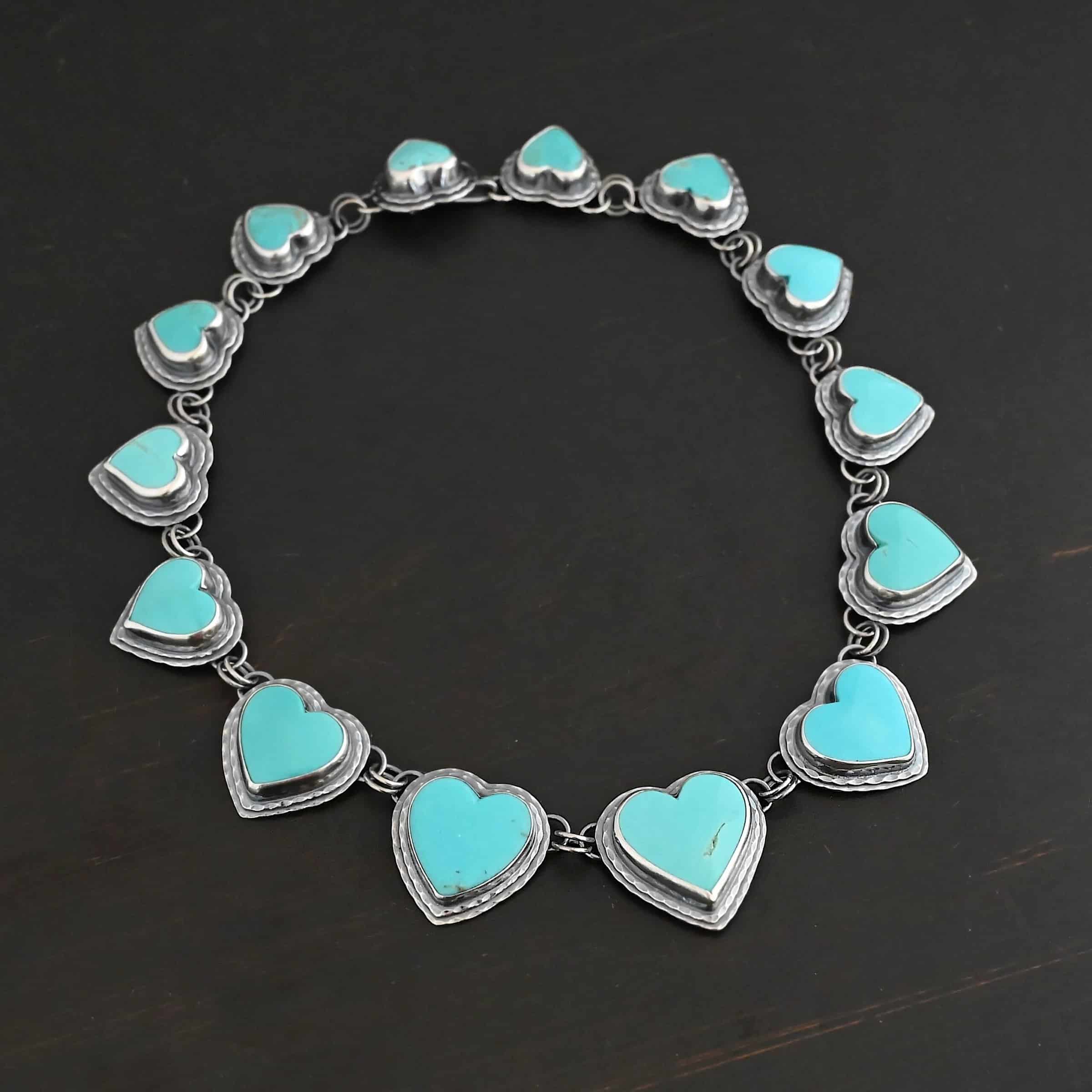 Heart & Soul Necklace Close up of the "Heart & Soul" Necklace. Made out of sterling silver and turquoise. There are fourteen turquoise hearts that circle around and create a collar necklace. Each turquoise heart is a light teal color and is set in sterling silver. Necklace is laying against a dark background.