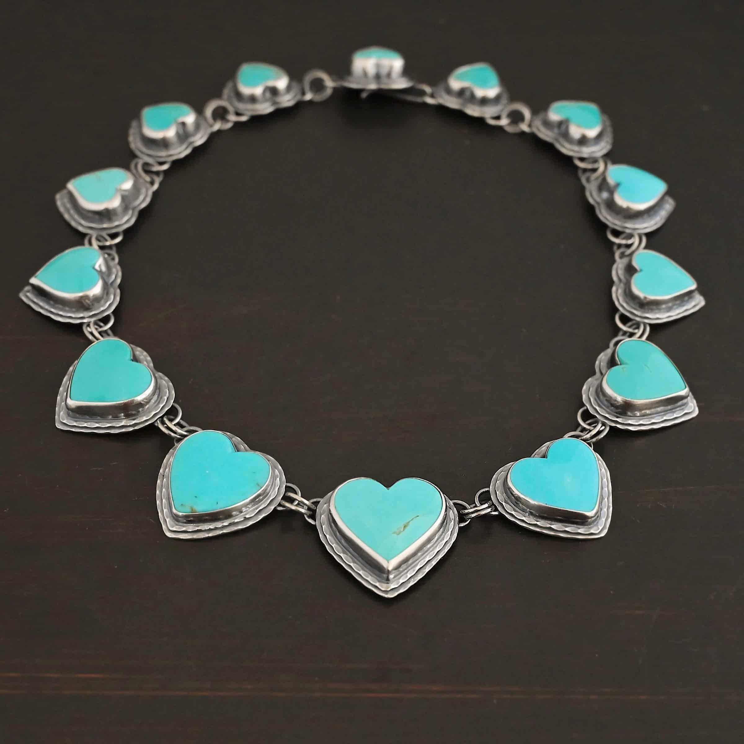 Heart & Soul Necklace Close up of the "Heart & Soul" Necklace. Made out of sterling silver and turquoise. There are fourteen turquoise hearts that circle around and create a collar necklace. Each turquoise heart is a light teal color and is set in sterling silver. Necklace is laying against a dark background.