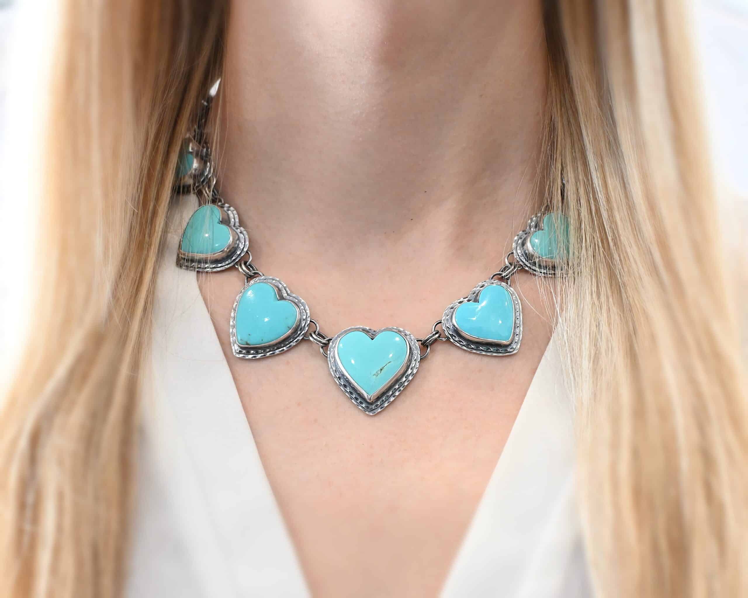 Heat & Soul Necklace Close up of the "Heart & Soul" Necklace. Made out of sterling silver and turquoise. There are fourteen turquoise hearts that circle around and create a collar necklace. Each turquoise heart is a light teal color and is set in sterling silver. Necklace is worn by a model at 16 inches long to demonstrate length. Model is wearing a white v-neck shirt.