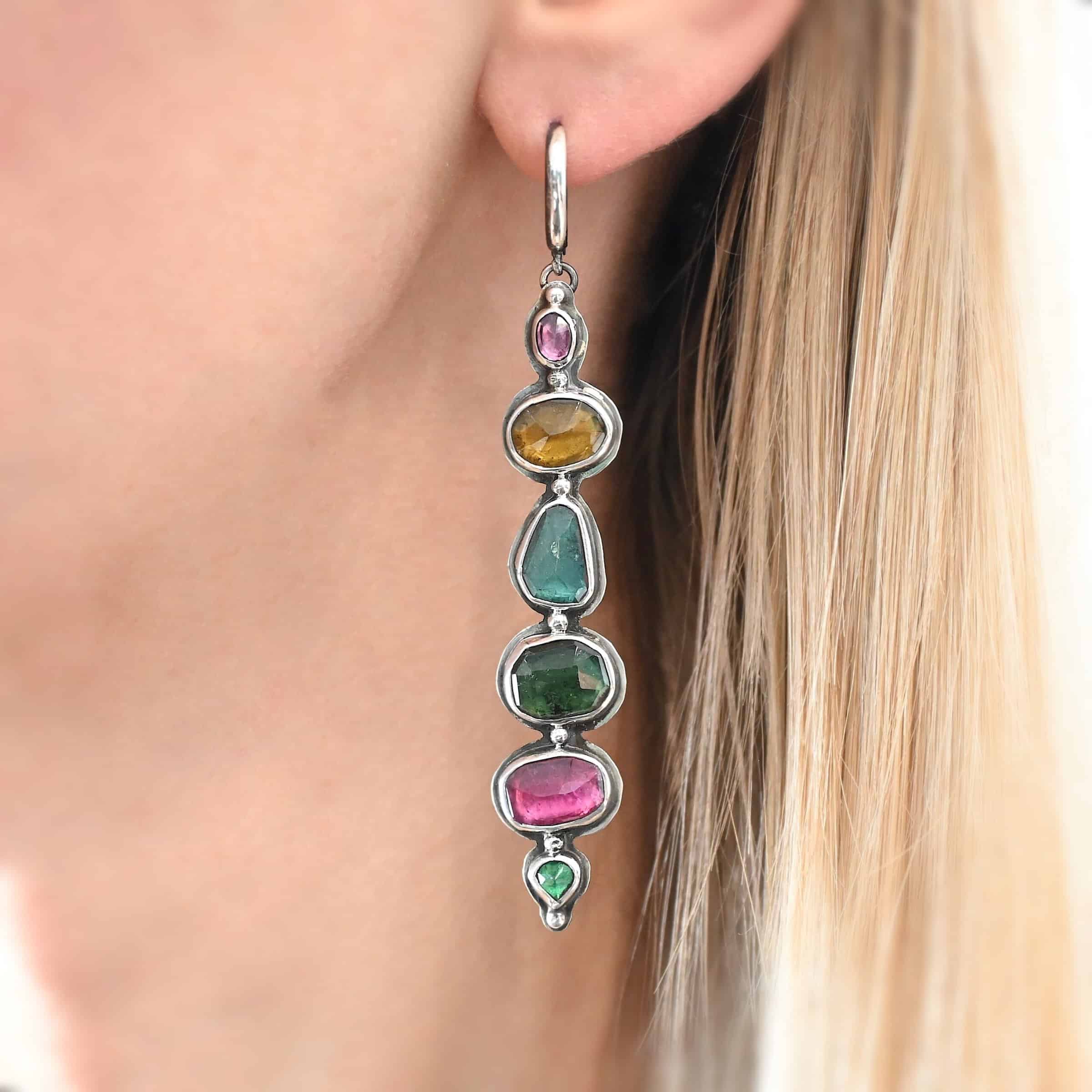 Color of Love Earrings Close up of the "Color Of Love" Earrings. Made out of sterling silver and sapphires. There are six sapphires, each a different shape, size, and color. There are two pink sapphires, two green, one blue, and one yellow. There are seven silver dots on the earring, one on the top of the first stone, five separating each stone afterwards, and one on the bottom of the final stone. Earring is worn by a model to demonstrate length and size of the earring.