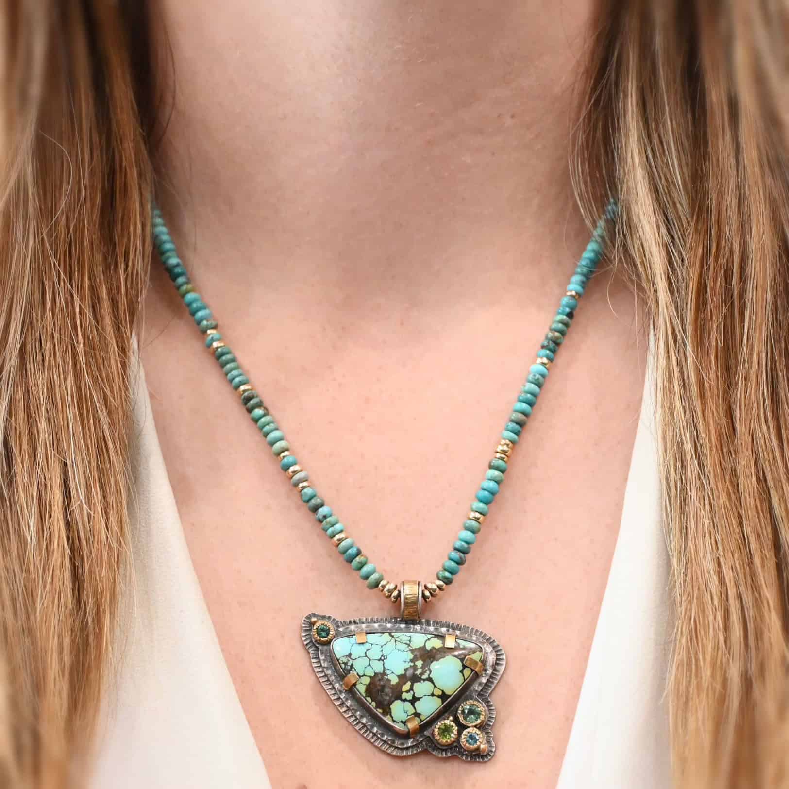 The "Trinity" Necklace as shown on a model. Made out of sterling silver, gold, turquoise, and tourmaline. The turquoise is the center stone, and the largest. It is triangular shaped. It is set in sterling silver, but has gold settings keeping it in place. There are three tourmalines on the bottom right side of the necklace, two green and one blue. There is another tourmaline in the top left corner of the necklace, also green. The chain for the necklace is turquoise and gold beads. The turquoise pendant is connected by a large gold link as well. Model is wearing a white v-neck.