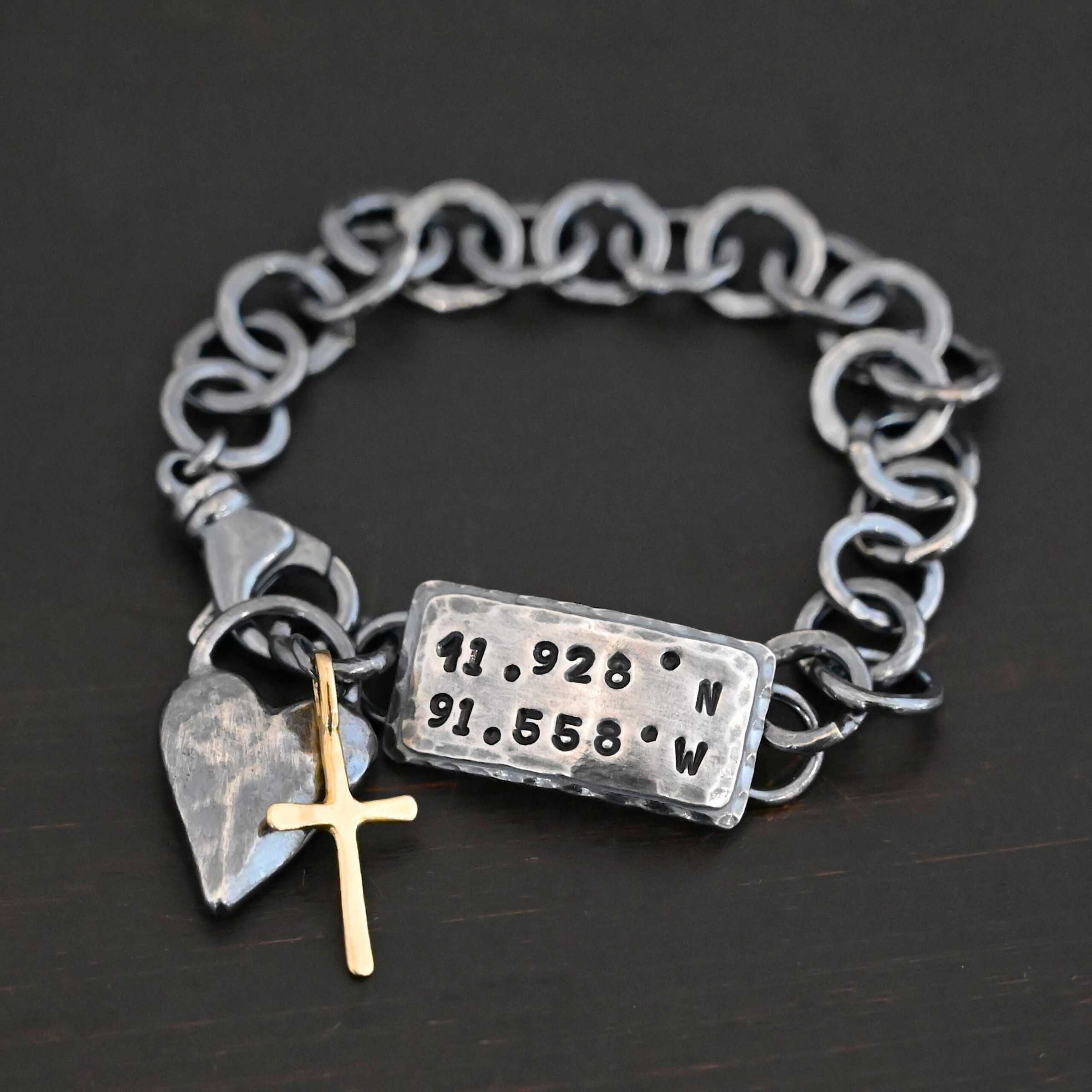 Home Sweet Home Bracelet Close up of the "Home Sweet Home" Bracelet. Made out of sterling silver and gold. There is a solid sterling silver heart with a gold cross. Both have loops on them and are attached to the link of a bracelet. The links are thick, hammered, circles. There is a large hammered rectangle tag on the bracelet as well. It has the coordinates 41.928.N and 91.558.W on it. The rectangle tag has two layer of sterling silver, the bottom of which is hammered. Bracelet is oxidized and laying against a dark background.