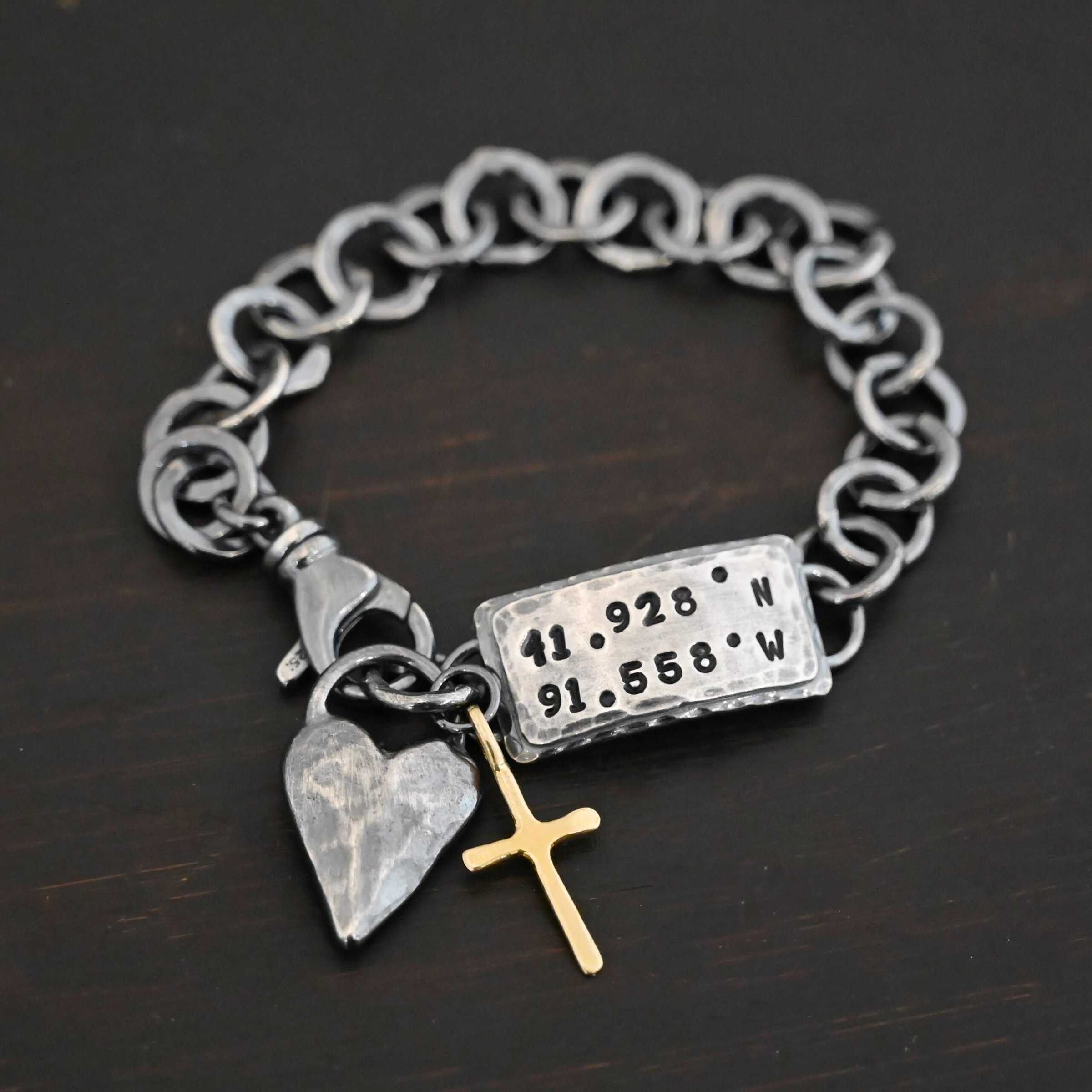 Home Sweet Home Bracelet Close up of the "Home Sweet Home" Bracelet. Made out of sterling silver and gold. There is a solid sterling silver heart with a gold cross. Both have loops on them and are attached to the link of a bracelet. The links are thick, hammered, circles. There is a large hammered rectangle tag on the bracelet as well. It has the coordinates 41.928.N and 91.558.W on it. The rectangle tag has two layer of sterling silver, the bottom of which is hammered. Bracelet is oxidized and laying against a dark background.
