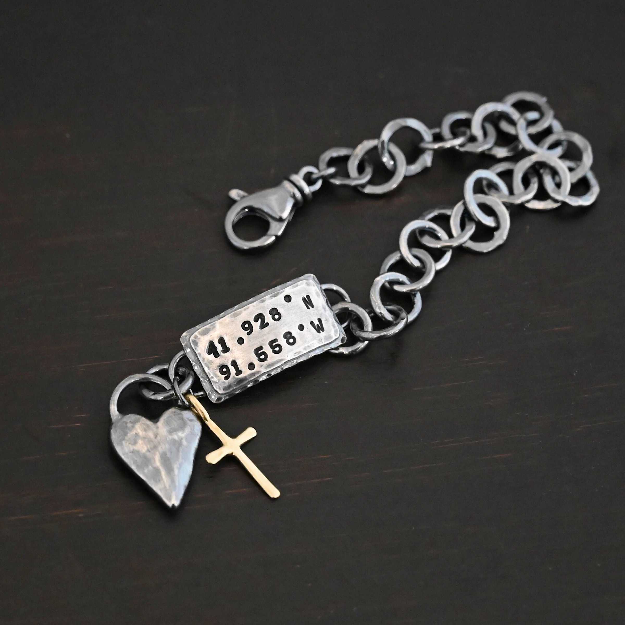 Home Sweet Home Bracelet Close up of the "Home Sweet Home" Bracelet. Made out of sterling silver and gold. There is a solid sterling silver heart with a gold cross. Both have loops on them and are attached to the link of a bracelet. The links are thick, hammered, circles. There is a large hammered rectangle tag on the bracelet as well. It has the coordinates 41.928.N and 91.558.W on it. The rectangle tag has two layer of sterling silver, the bottom of which is hammered. Bracelet is oxidized and laying against a dark background.