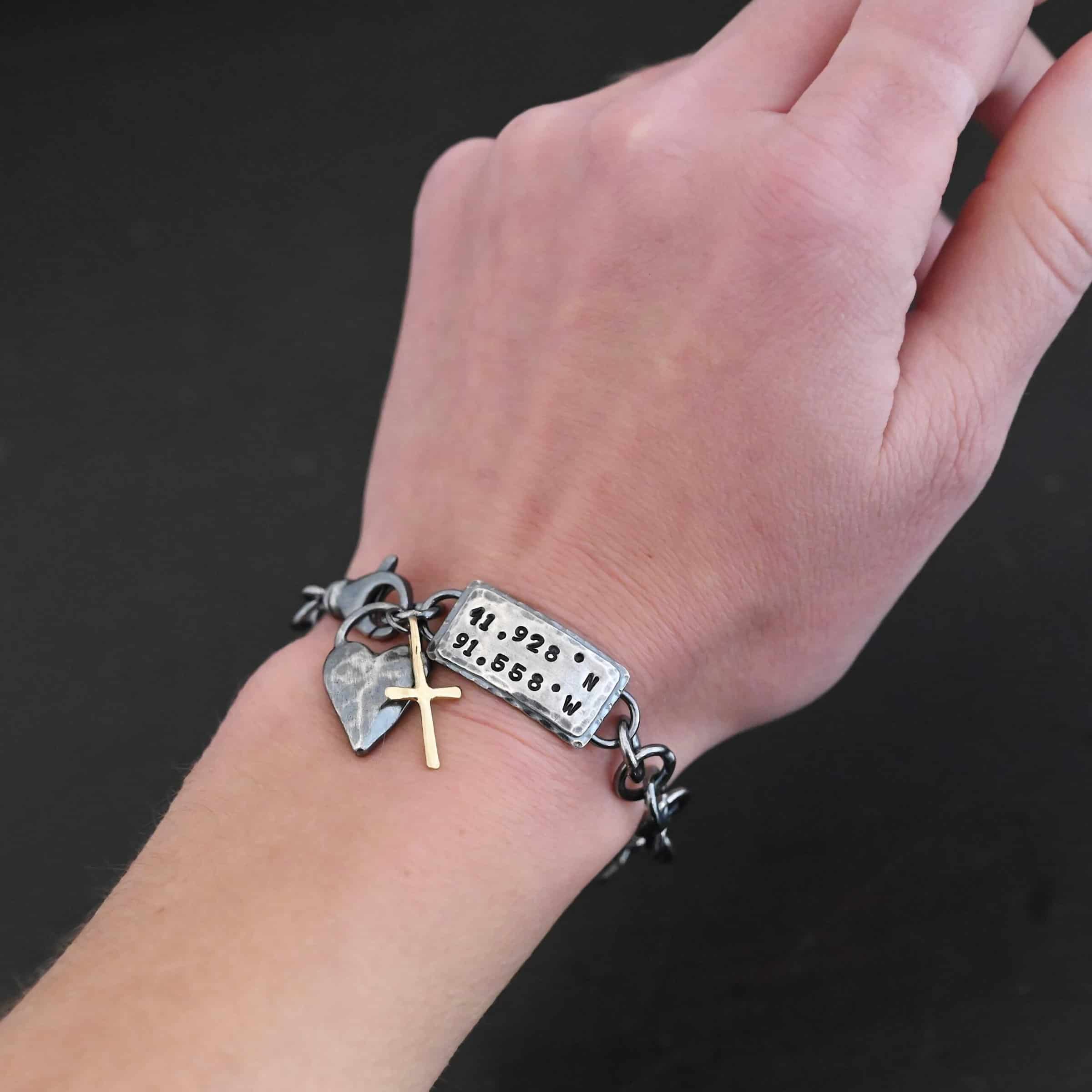 Home Sweet Home Bracelet Close up of the "Home Sweet Home" Bracelet. Made out of sterling silver and gold. There is a solid sterling silver heart with a gold cross. Both have loops on them and are attached to the link of a bracelet. The links are thick, hammered, circles. There is a large hammered rectangle tag on the bracelet as well. It has the coordinates 41.928.N and 91.558.W on it. The rectangle tag has two layer of sterling silver, the bottom of which is hammered. Bracelet is oxidized worn on a models wrist to demonstrate size.