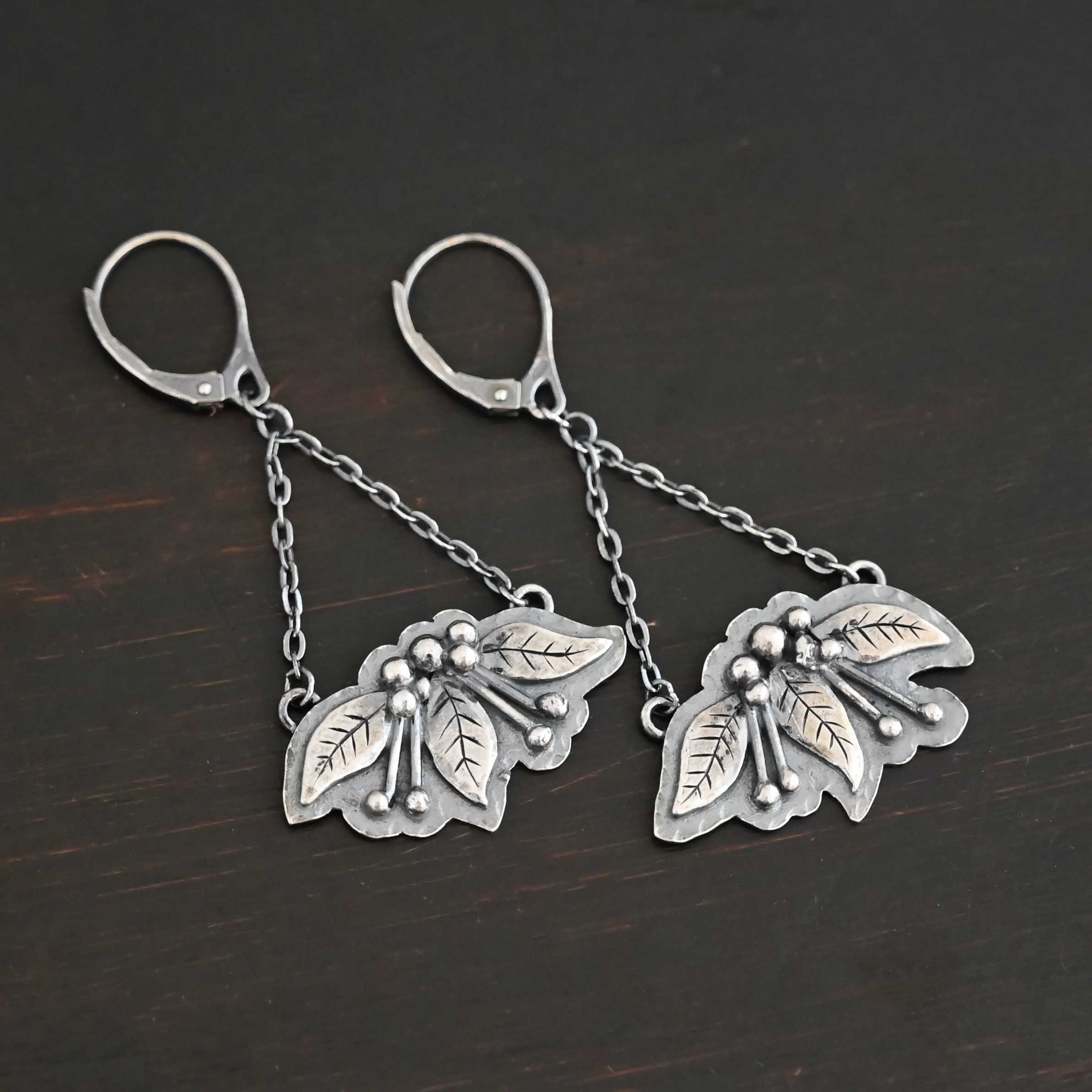 Close up of the "Paisley" Earrings. Made out of sterling silver. There are three silver leaves for each earring. Each earring also has five silver balls that separate each leaf on the top and four more silver balls on each side of the middle leaf with lines of silver leading to the top balls. On the back of each earring are two loops, one on each end of the earring. Attached to each loop is a strand of sterling silver. The strands meet to form a triangle shape, at which the top of the triangle is the leverback ear wire. Earrings are set against a dark background.