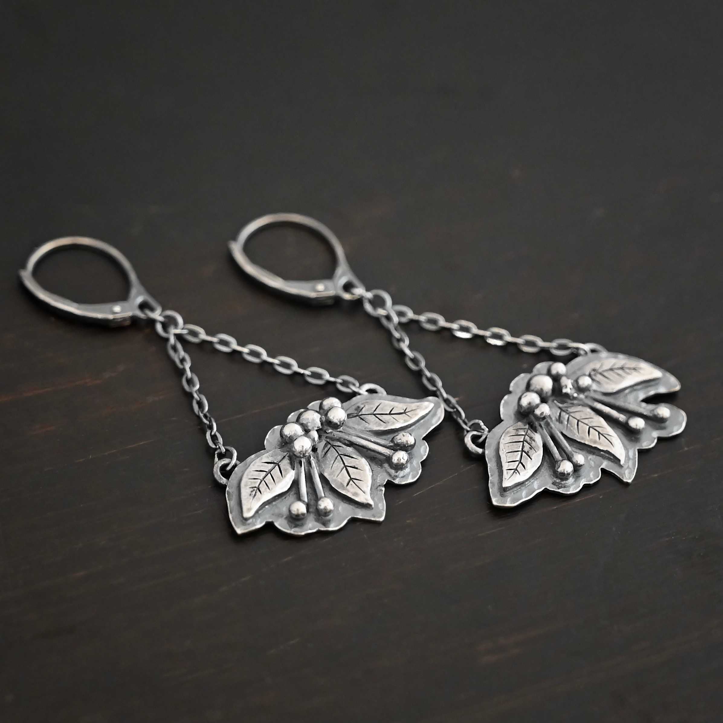 Close up of the "Paisley" Earrings. Made out of sterling silver. There are three silver leaves for each earring. Each earring also has five silver balls that separate each leaf on the top and four more silver balls on each side of the middle leaf with lines of silver leading to the top balls. On the back of each earring are two loops, one on each end of the earring. Attached to each loop is a strand of sterling silver. The strands meet to form a triangle shape, at which the top of the triangle is the leverback ear wire. Earrings are set against a dark background.