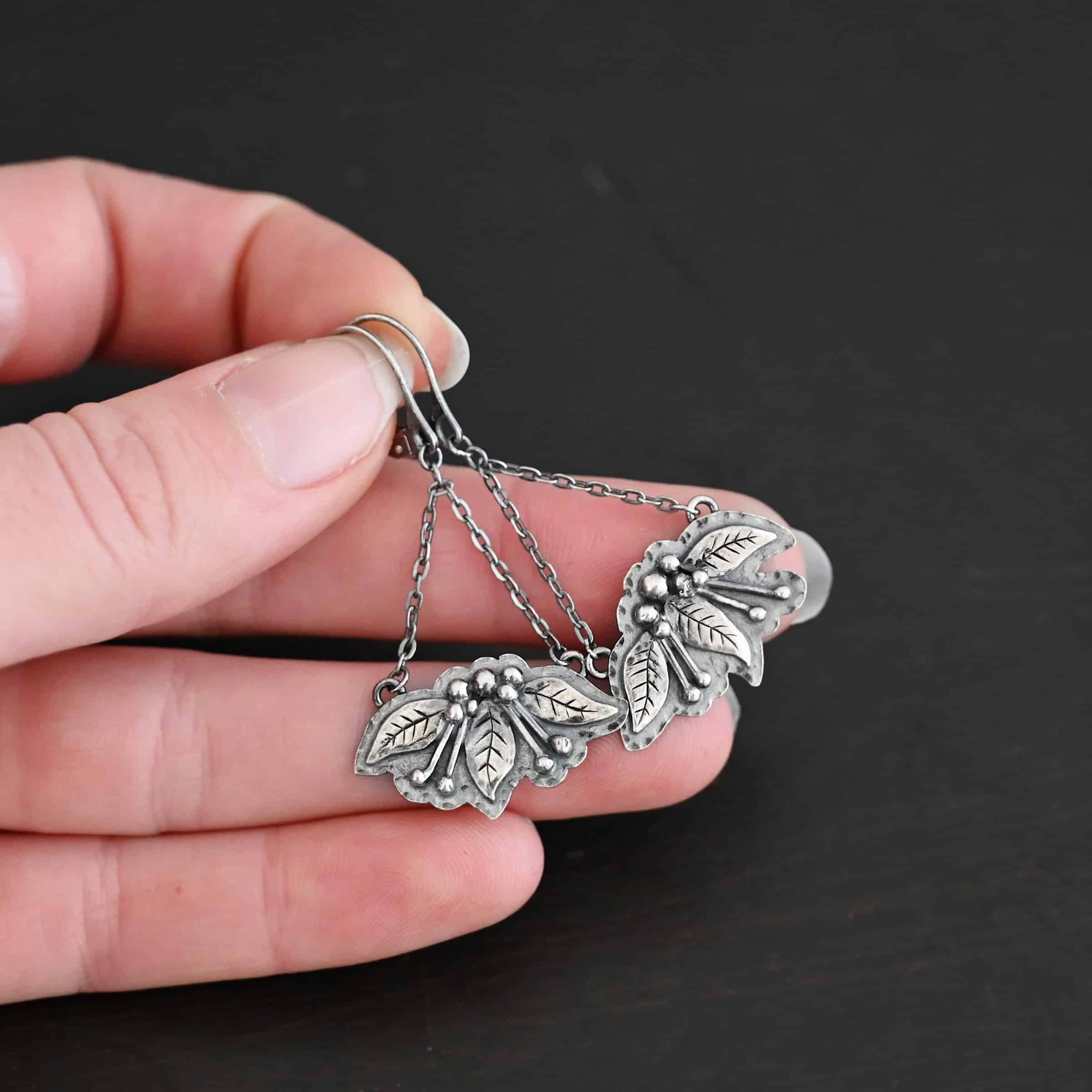 Close up of the "Paisley" Earrings. Made out of sterling silver. There are three silver leaves for each earring. Each earring also has five silver balls that separate each leaf on the top and four more silver balls on each side of the middle leaf with lines of silver leading to the top balls. On the back of each earring are two loops, one on each end of the earring. Attached to each loop is a strand of sterling silver. The strands meet to form a triangle shape, at which the top of the triangle is the leverback ear wire. Earrings are held in the fingertips of a hand to demonstrate size.