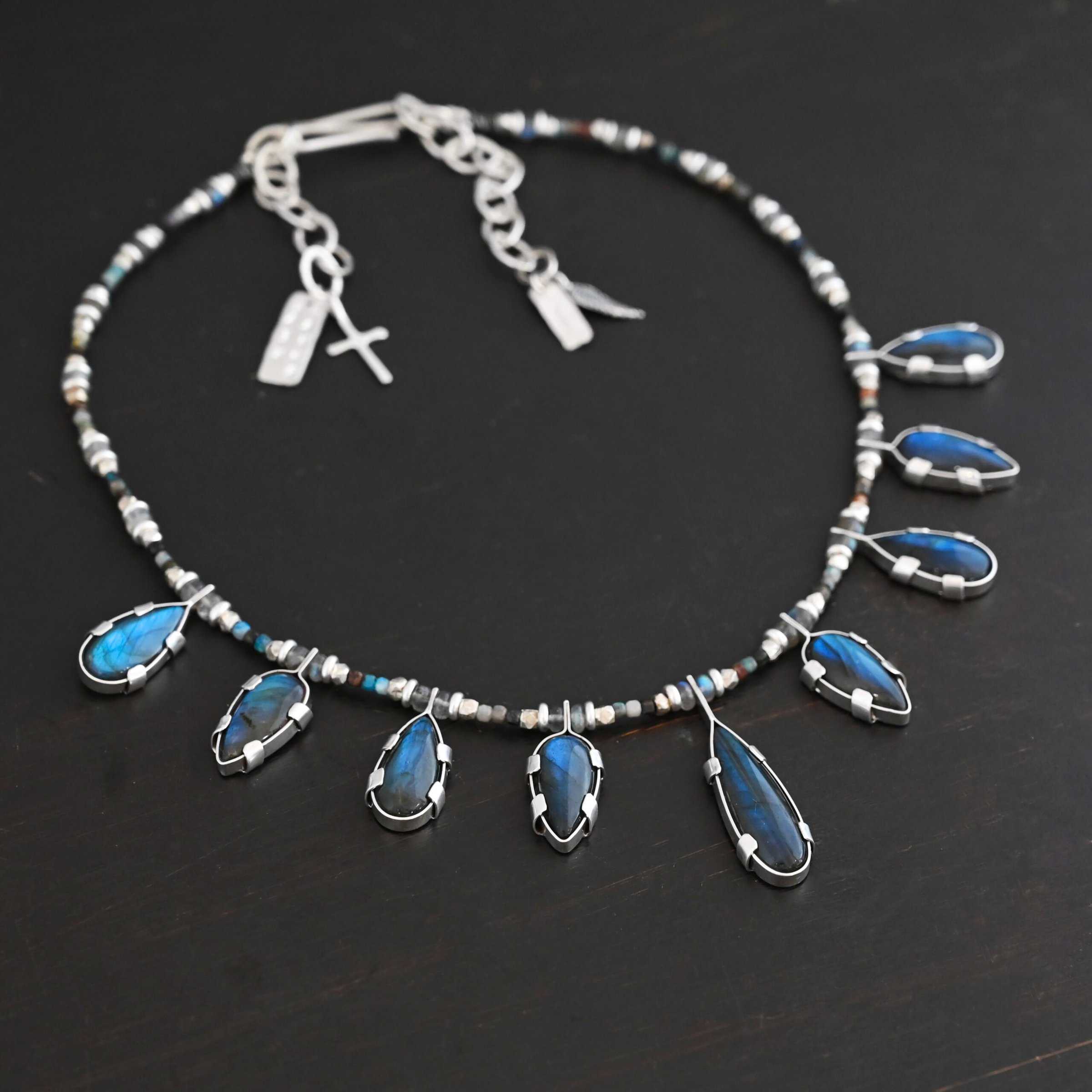 Rain Dance Necklace Close up of the "Rain Dance" Necklace. Made out of sterling silver and labradorite, with labradorite beads. There are eight teardrop shaped labradorites, and each stone alternates from point facing upwards to point facing downwards. Each one is connected to a chain that is made out of labradorite and chrysocolla beads. Each labradorite has four tabs, two on each side of the stone, that are clamped down on the edges of it to keep it held in. Necklace is shown against a dark background.