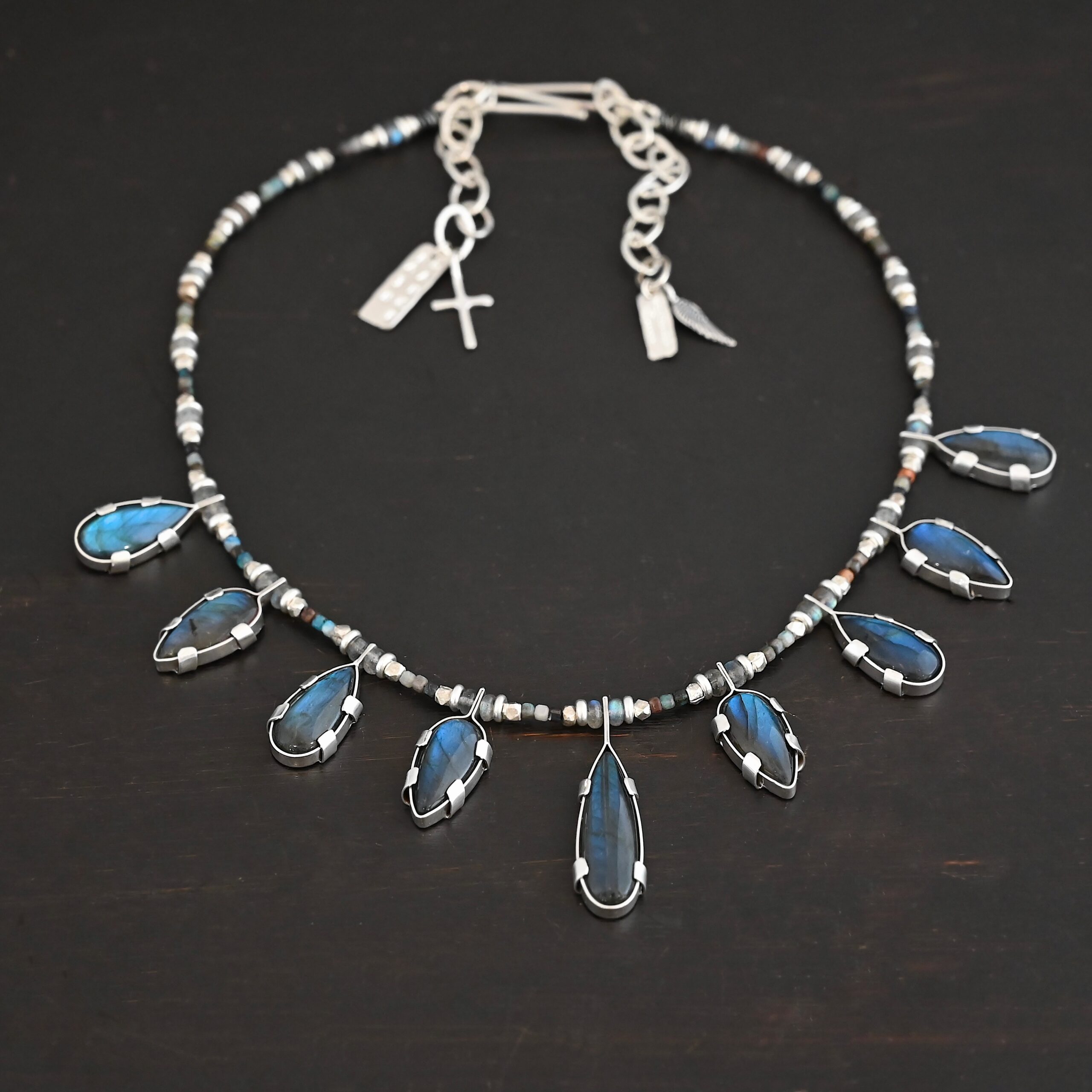 Rain Dance Necklace Close up of the "Rain Dance" Necklace. Made out of sterling silver and labradorite, with labradorite beads. There are eight teardrop shaped labradorites, and each stone alternates from point facing upwards to point facing downwards. Each one is connected to a chain that is made out of labradorite and chrysocolla beads. Each labradorite has four tabs, two on each side of the stone, that are clamped down on the edges of it to keep it held in. Necklace is shown against a dark background.