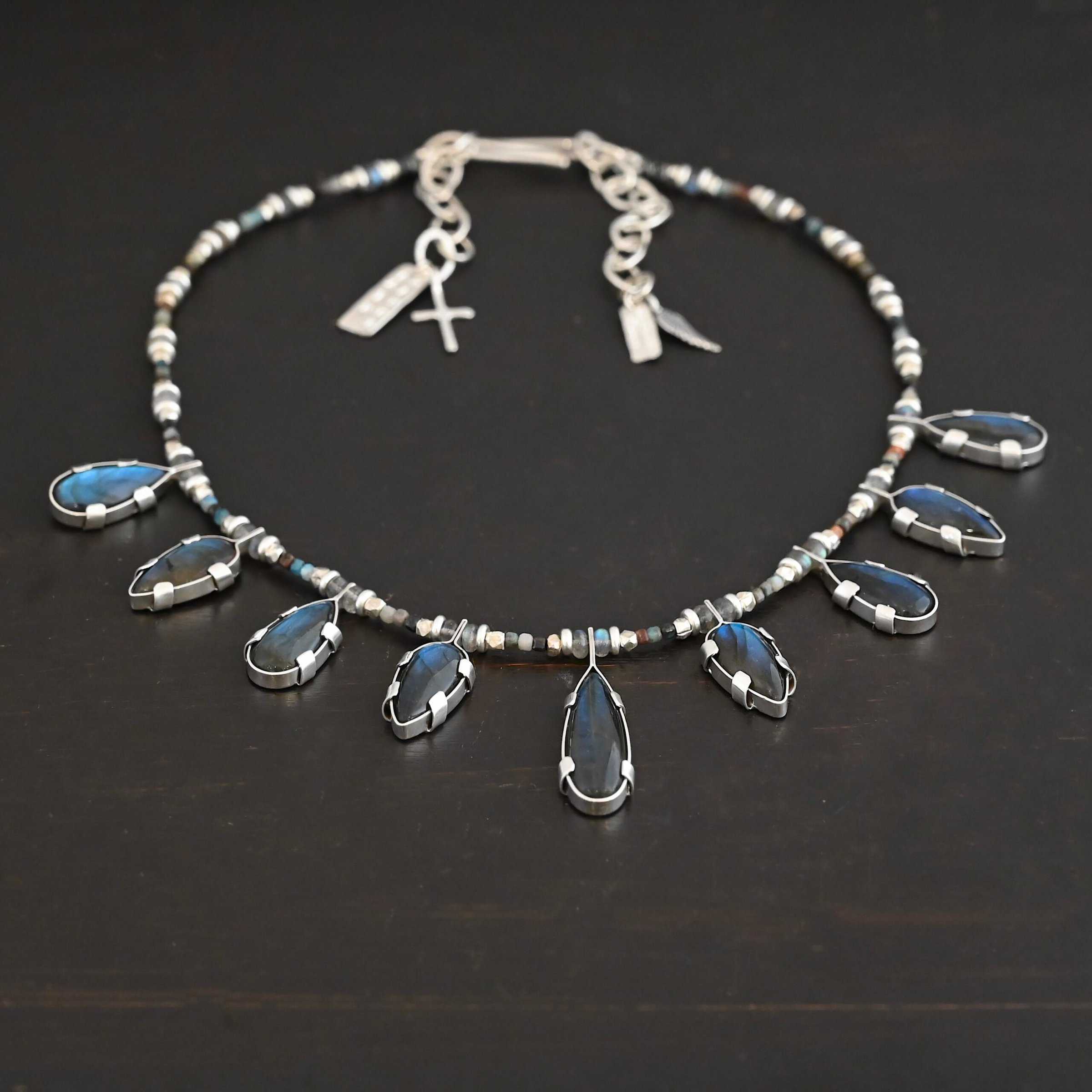 Rain Dance Necklace Close up of the "Rain Dance" Necklace. Made out of sterling silver and labradorite, with labradorite beads. There are eight teardrop shaped labradorites, and each stone alternates from point facing upwards to point facing downwards. Each one is connected to a chain that is made out of labradorite and chrysocolla beads. Each labradorite has four tabs, two on each side of the stone, that are clamped down on the edges of it to keep it held in. Necklace is shown against a dark background.