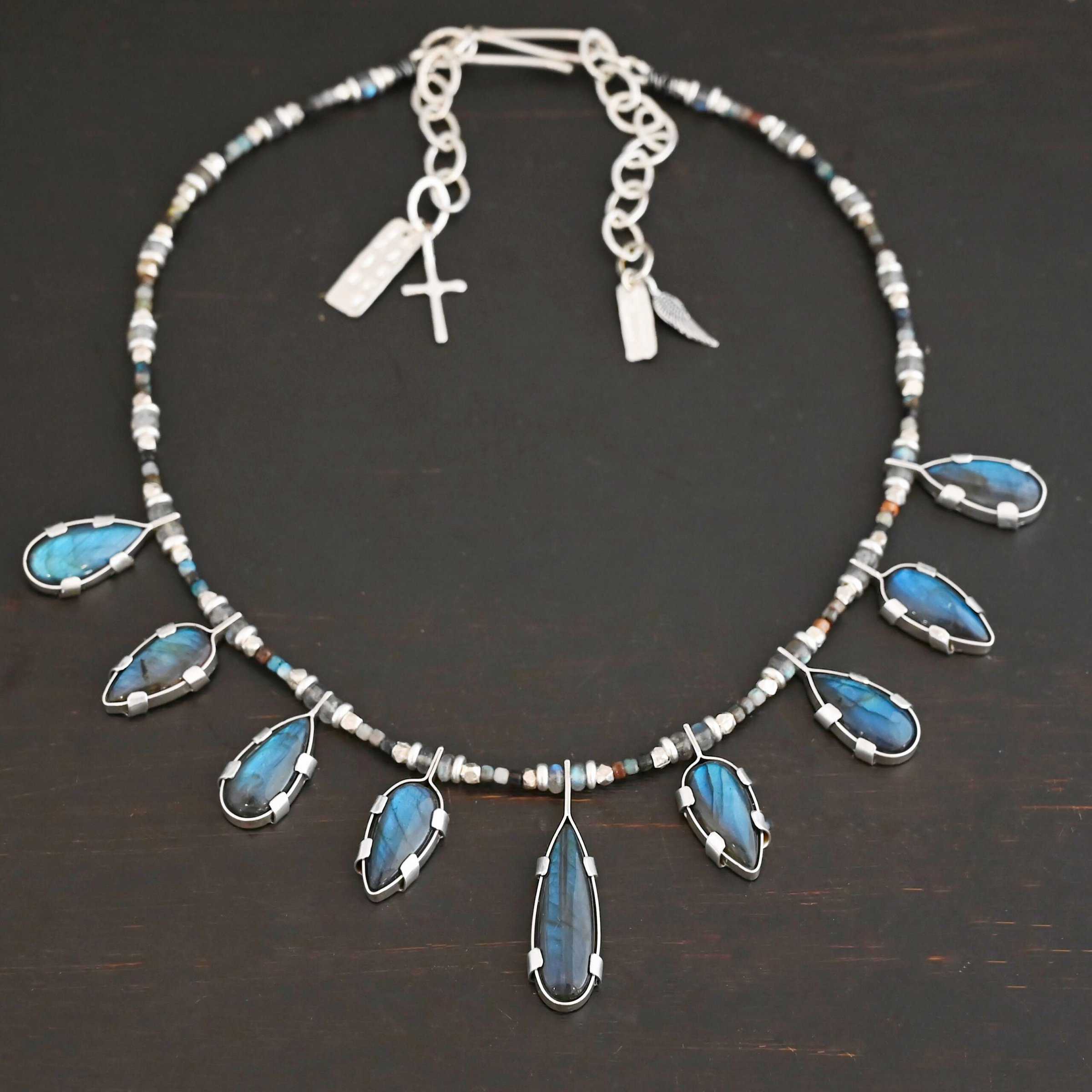 Rain Dance Necklace Close up of the "Rain Dance" Necklace. Made out of sterling silver and labradorite, with labradorite beads. There are eight teardrop shaped labradorites, and each stone alternates from point facing upwards to point facing downwards. Each one is connected to a chain that is made out of labradorite and chrysocolla beads. Each labradorite has four tabs, two on each side of the stone, that are clamped down on the edges of it to keep it held in. Necklace is shown against a dark background.
