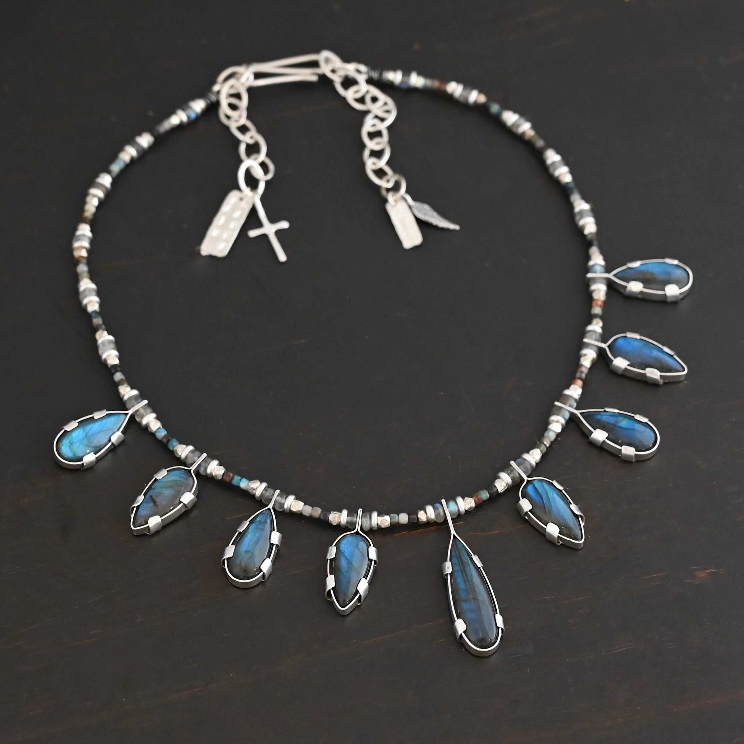 Rain Dance Necklace Close up of the "Rain Dance" Necklace. Made out of sterling silver and labradorite, with labradorite beads. There are eight teardrop shaped labradorites, and each stone alternates from point facing upwards to point facing downwards. Each one is connected to a chain that is made out of labradorite and chrysocolla beads. Each labradorite has four tabs, two on each side of the stone, that are clamped down on the edges of it to keep it held in. Necklace is shown against a dark background.