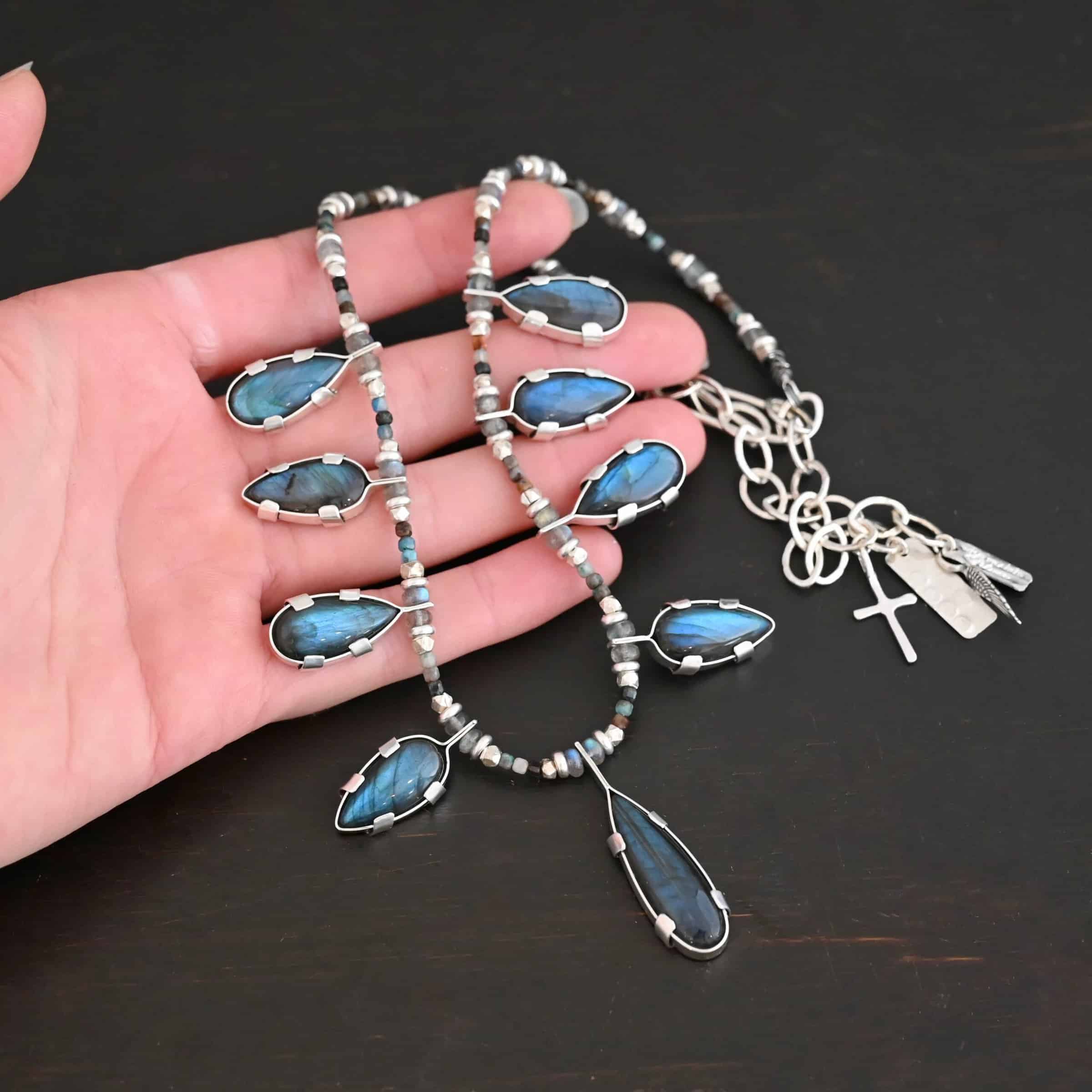 Rain Dance Necklace Close up of the "Rain Dance" Necklace. Made out of sterling silver and labradorite, with labradorite beads. There are eight teardrop shaped labradorites, and each stone alternates from point facing upwards to point facing downwards. Each one is connected to a chain that is made out of labradorite and chrysocolla beads. Each labradorite has four tabs, two on each side of the stone, that are clamped down on the edges of it to keep it held in. Necklace is shown against the fingertips of a hand to demonstrate size.