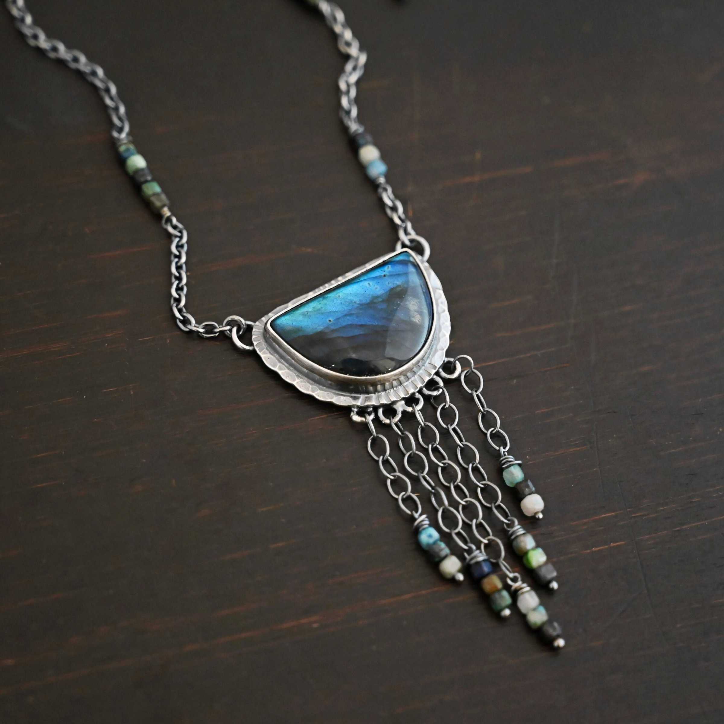 Dream Catcher Necklace Close up of the "Dream Catcher" Necklace. Made out of sterling silver, labradorite, and chrysocolla beads. The labradorite is a half circle shape, with the flat half at the top from where the circle would be cut in half. There are five short strands of silver chain with chrysocolla beads on each end of the chains. The labradorite is set in hammered sterling silver and on each end of the top of the setting is a loop. Attached to the loops are silver chain with chrysocolla beads on them. Necklace is laying against a dark background.