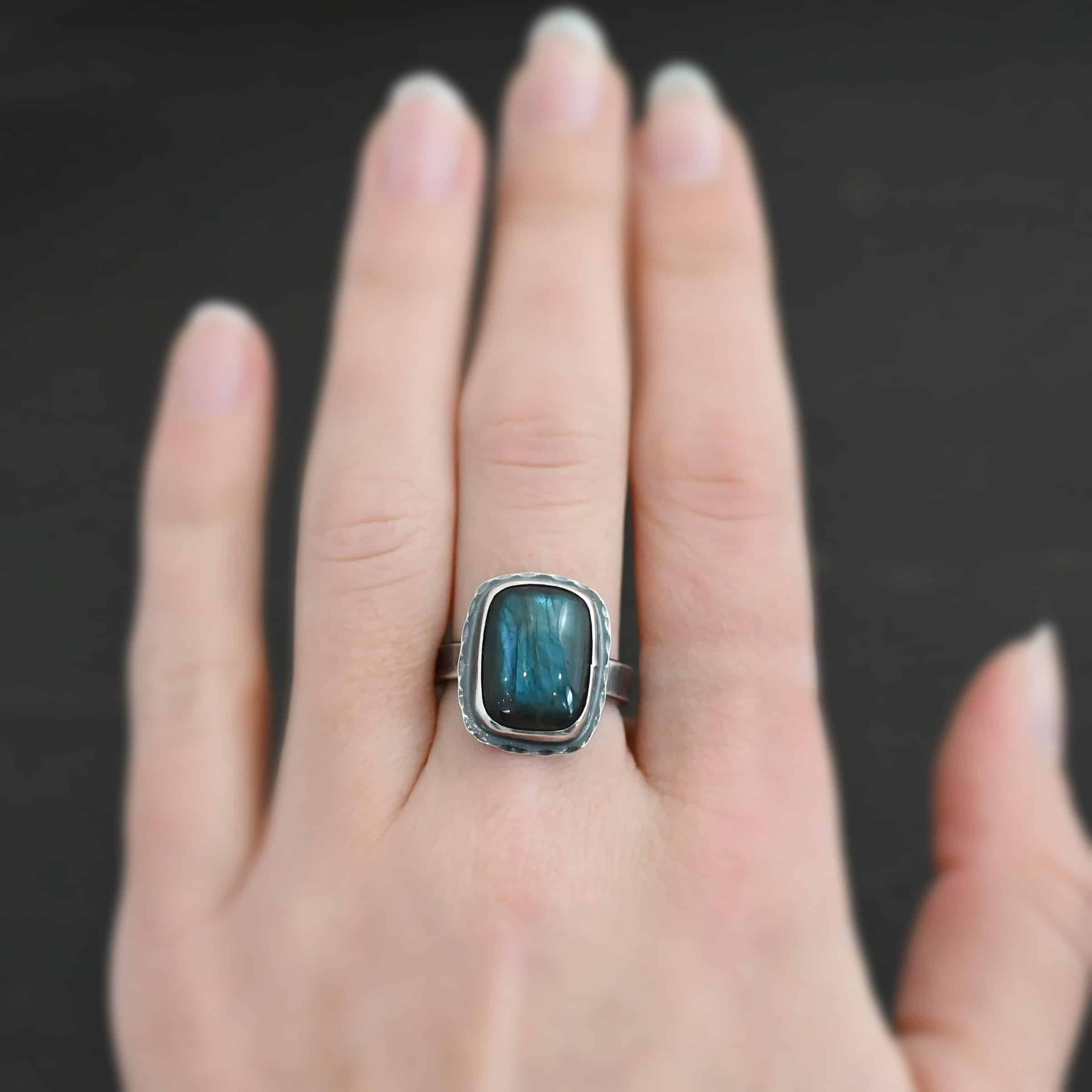 Close up of the "Rainstorm" Ring. Made out of sterling silver and labradorite. The labradorite is a square shape and a darker blue color with light blues scattered throughout it. The stone is set in sterling silver with hammered sterling silver surrounding it. Ring is worn on a models middle finger to demonstrate size.