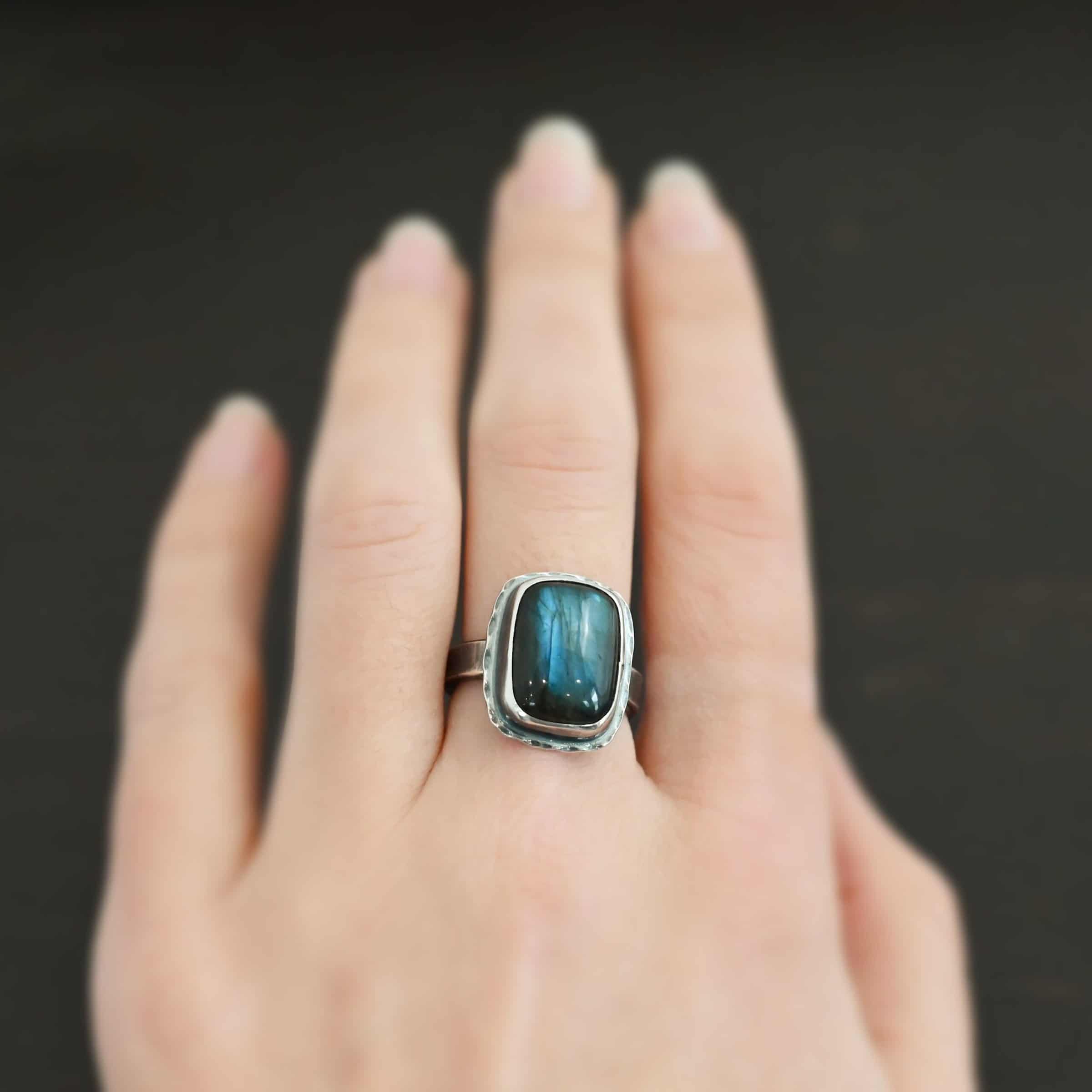 Close up of the "Rainstorm" Ring. Made out of sterling silver and labradorite. The labradorite is a square shape and a darker blue color with light blues scattered throughout it. The stone is set in sterling silver with hammered sterling silver surrounding it. Ring is worn on a models middle finger to demonstrate size.