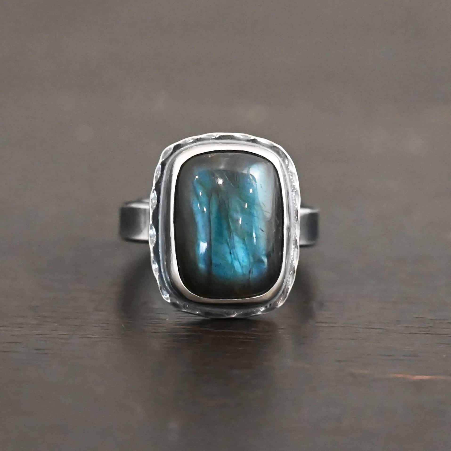 Close up of the "Rainstorm" Ring. Made out of sterling silver and labradorite. The labradorite is a square shape and a darker blue color with light blues scattered throughout it. The stone is set in sterling silver with hammered sterling silver surrounding it. Ring is set against a dark background.