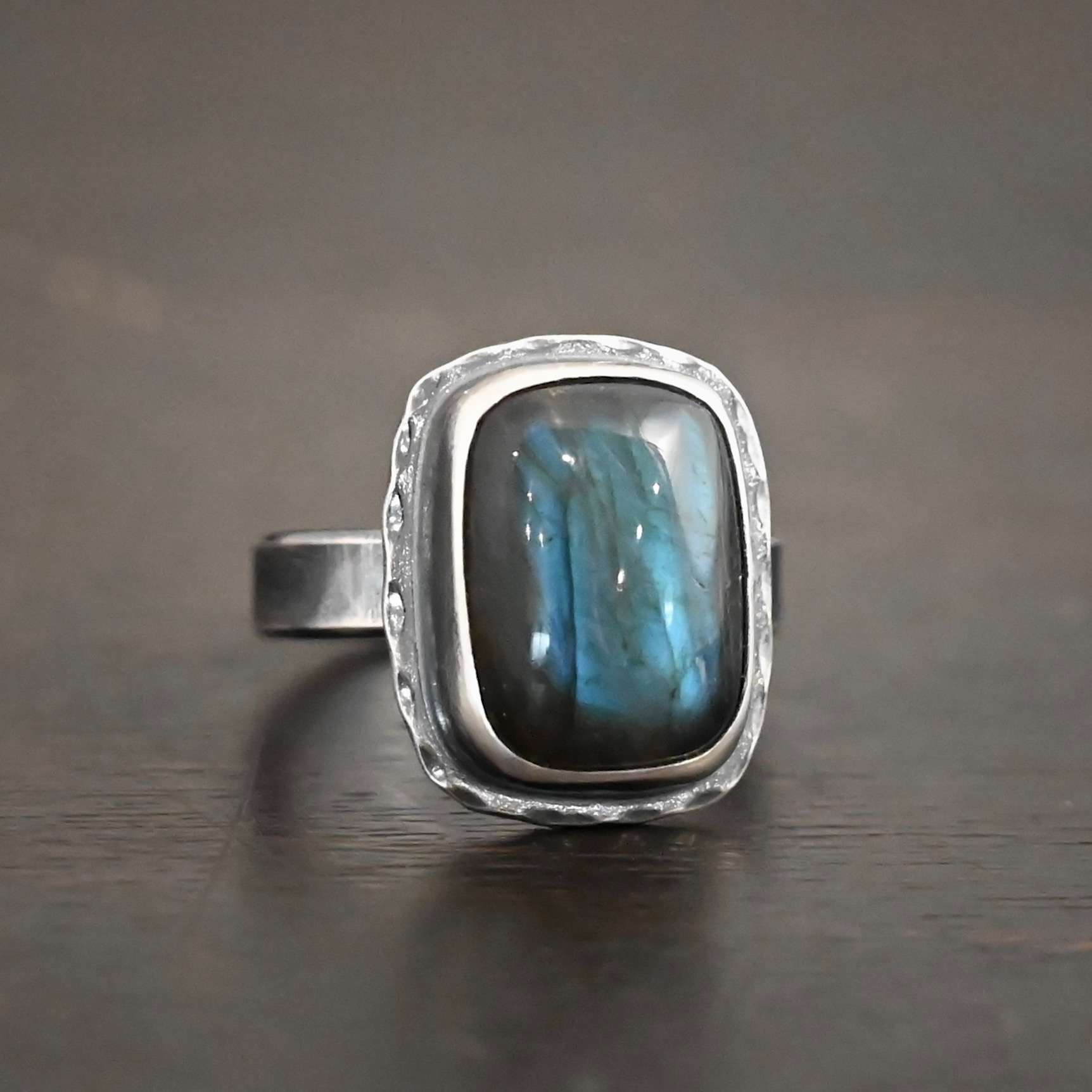 Close up of the "Rainstorm" Ring. Made out of sterling silver and labradorite. The labradorite is a square shape and a darker blue color with light blues scattered throughout it. The stone is set in sterling silver with hammered sterling silver surrounding it. Ring is set against a dark background.