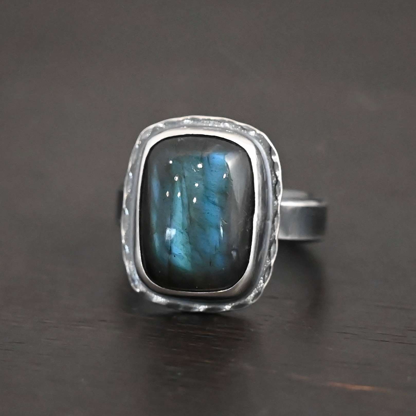 Close up of the "Rainstorm" Ring. Made out of sterling silver and labradorite. The labradorite is a square shape and a darker blue color with light blues scattered throughout it. The stone is set in sterling silver with hammered sterling silver surrounding it. Ring is set against a dark background.