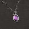 Close up of the "Thea" Necklace. Made out of sterling silver and amethyst. The amethyst is an oval shape. There are two small silver balls, one on the top of the oval and one on the bottom. The amethyst has hammered and oxidized sterling silver surrounding the outside of it. There is a loop on the back of the pendant that is attached to three jump rings. The jump rings are attached to the chain of the necklace. Necklace is laying against a dark background.