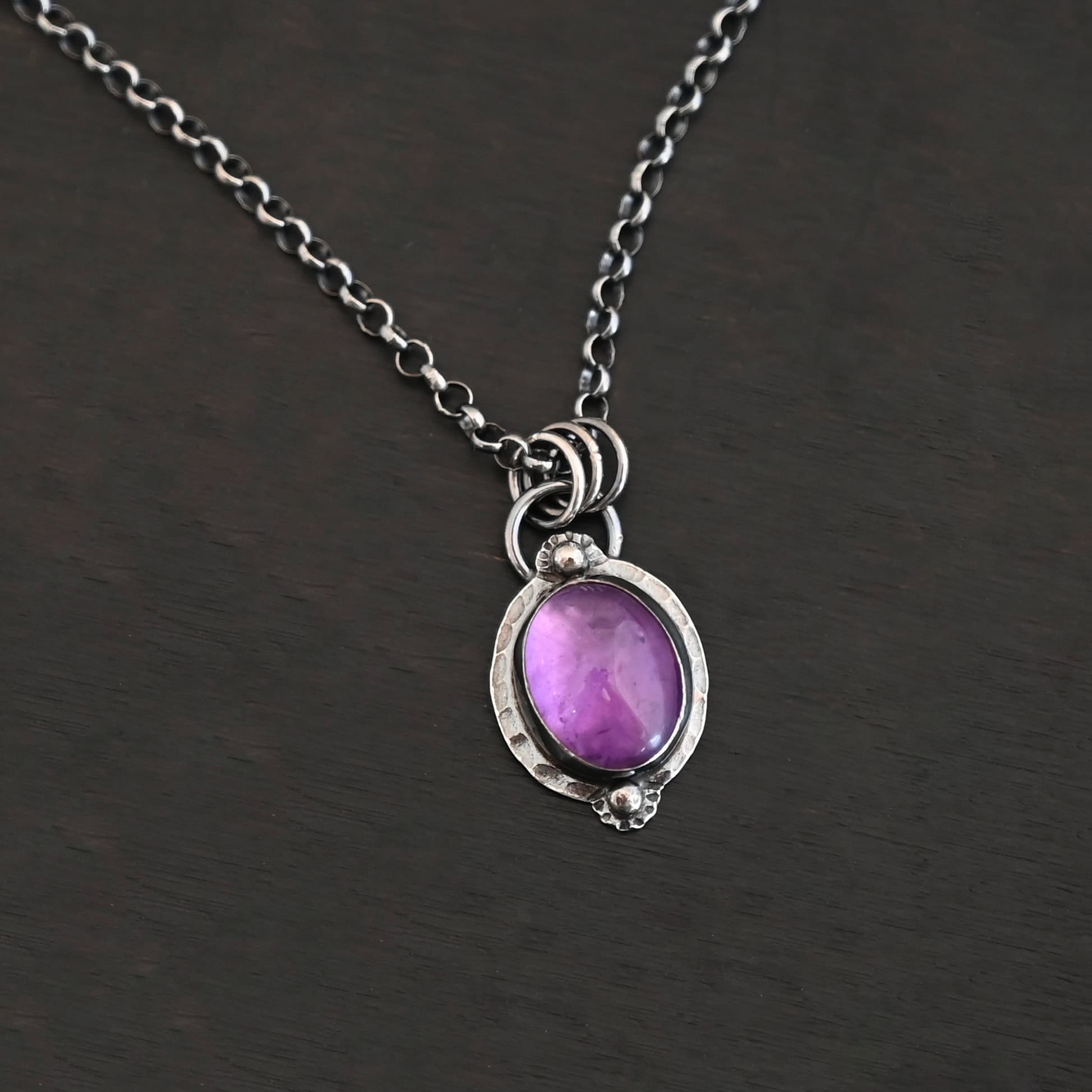 Close up of the "Thea" Necklace. Made out of sterling silver and amethyst. The amethyst is an oval shape. There are two small silver balls, one on the top of the oval and one on the bottom. The amethyst has hammered and oxidized sterling silver surrounding the outside of it. There is a loop on the back of the pendant that is attached to three jump rings. The jump rings are attached to the chain of the necklace. Necklace is laying against a dark background.