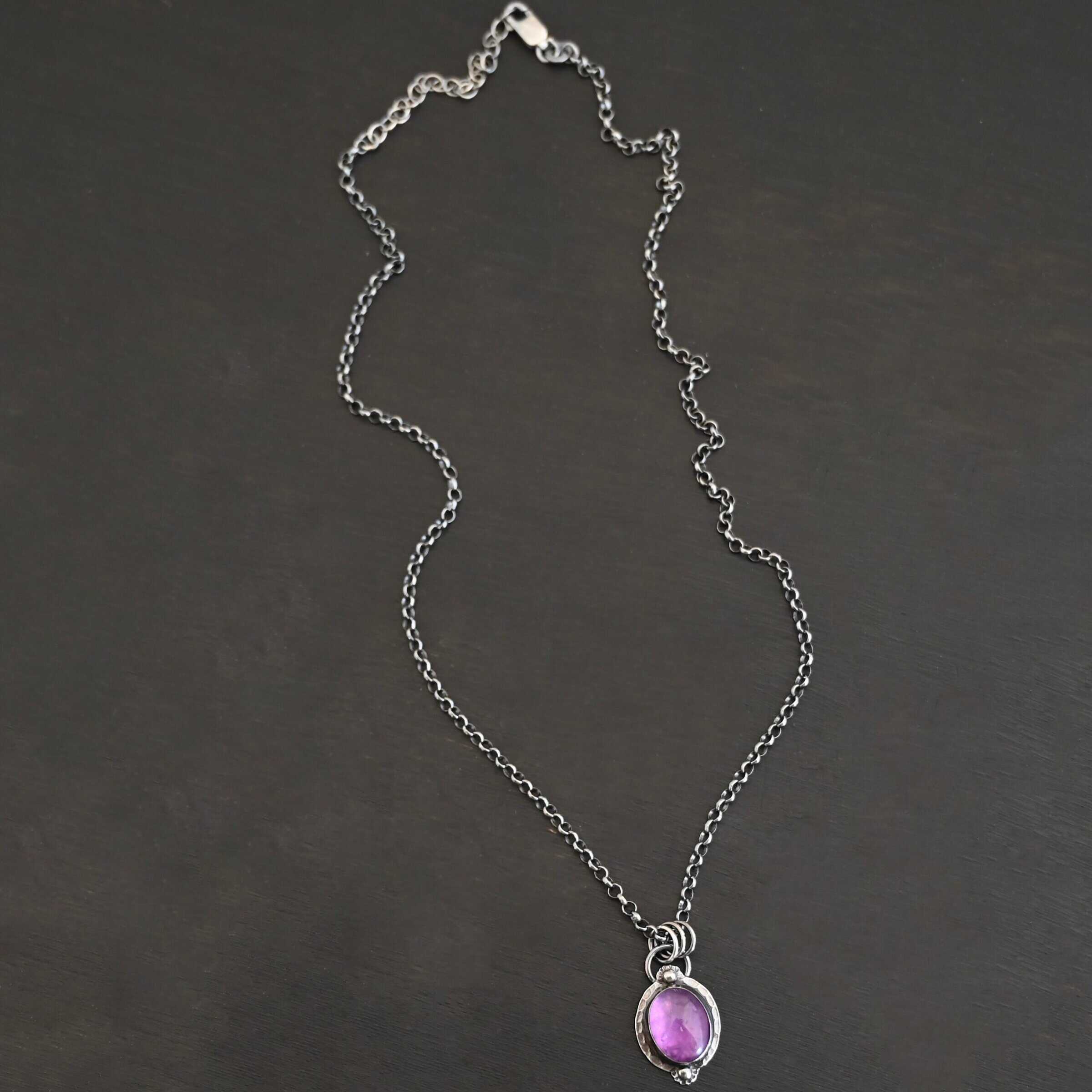 Close up of the "Thea" Necklace. Made out of sterling silver and amethyst. The amethyst is an oval shape. There are two small silver balls, one on the top of the oval and one on the bottom. The amethyst has hammered and oxidized sterling silver surrounding the outside of it. There is a loop on the back of the pendant that is attached to three jump rings. The jump rings are attached to the chain of the necklace. Necklace is laying against a dark background.