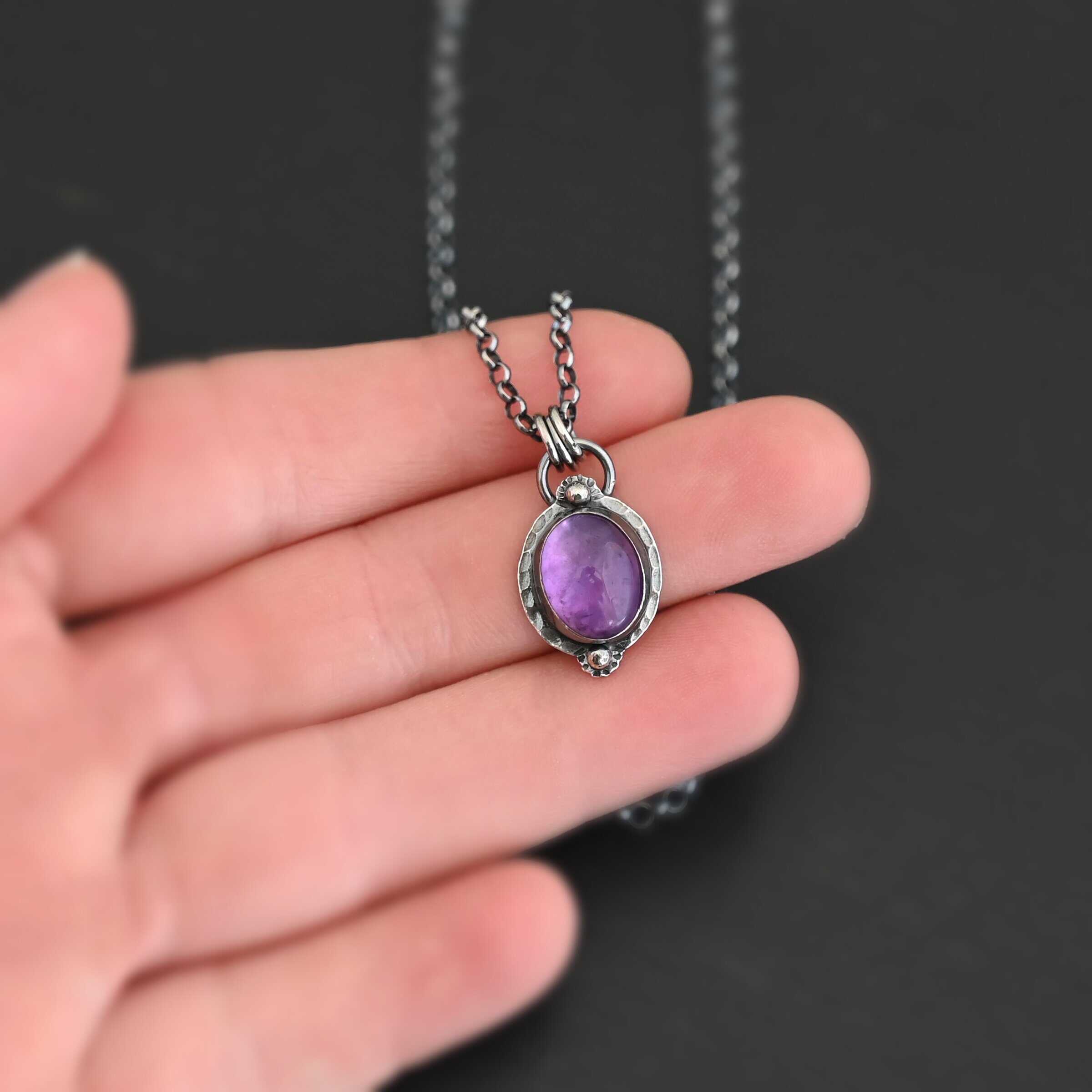 Close up of the "Thea" Necklace. Made out of sterling silver and amethyst. The amethyst is an oval shape. There are two small silver balls, one on the top of the oval and one on the bottom. The amethyst has hammered and oxidized sterling silver surrounding the outside of it. There is a loop on the back of the pendant that is attached to three jump rings. The jump rings are attached to the chain of the necklace.