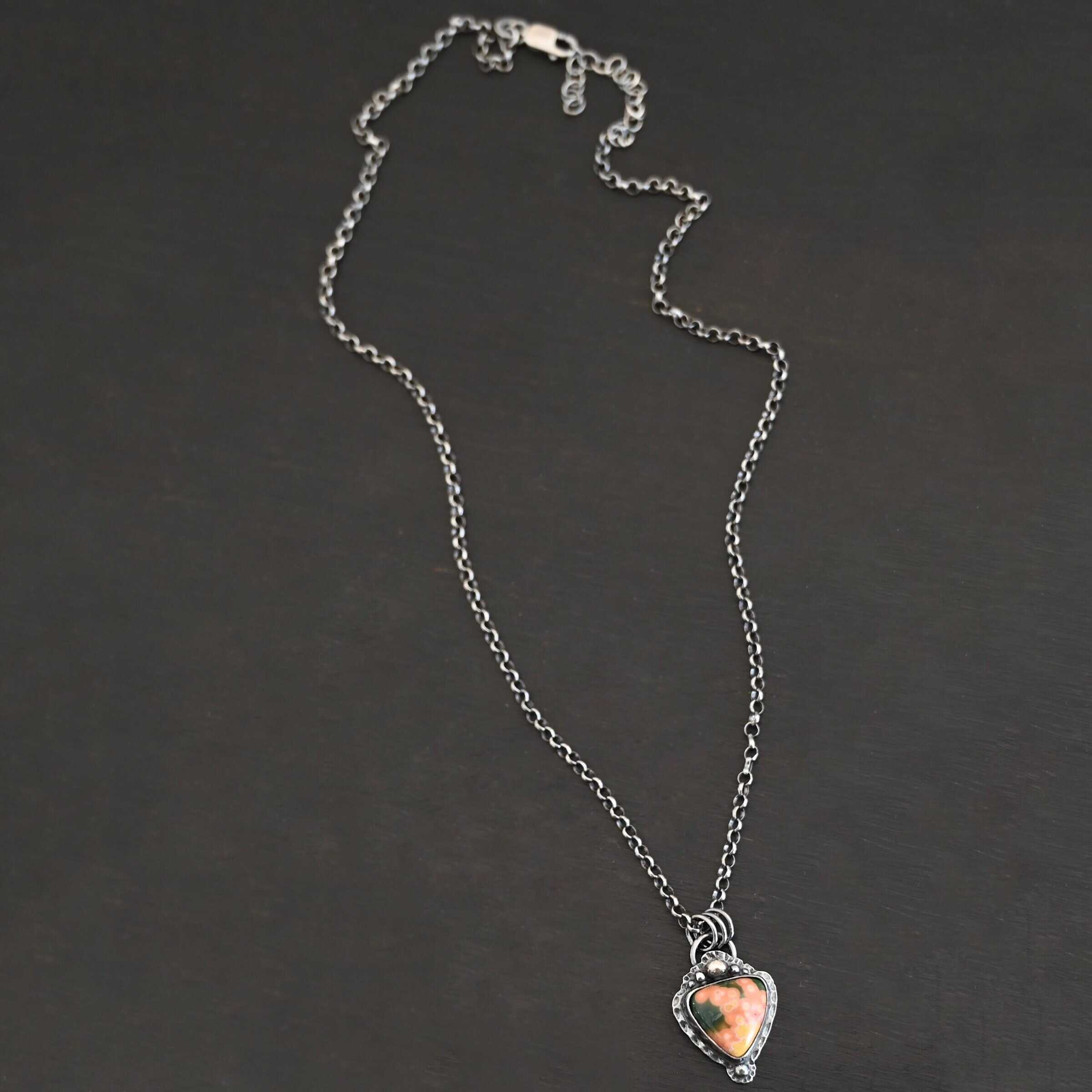Close up of the "Mesa" Necklace. Made out of sterling silver and ocean jasper. The jasper is a triangle shape, point facing downwards and is primarily orange and yellow with dark green scattered around the top of it. There are five silver dots on the top of the pendant which is flat since the top point of the triangle is facing downwards. There is a silver dot on the bottom of the tip of the necklace. There is a loop on the back of the pendant in which jump rings are attached to. There is a chain attached to the jump rings. Necklace is laying against a dark background.