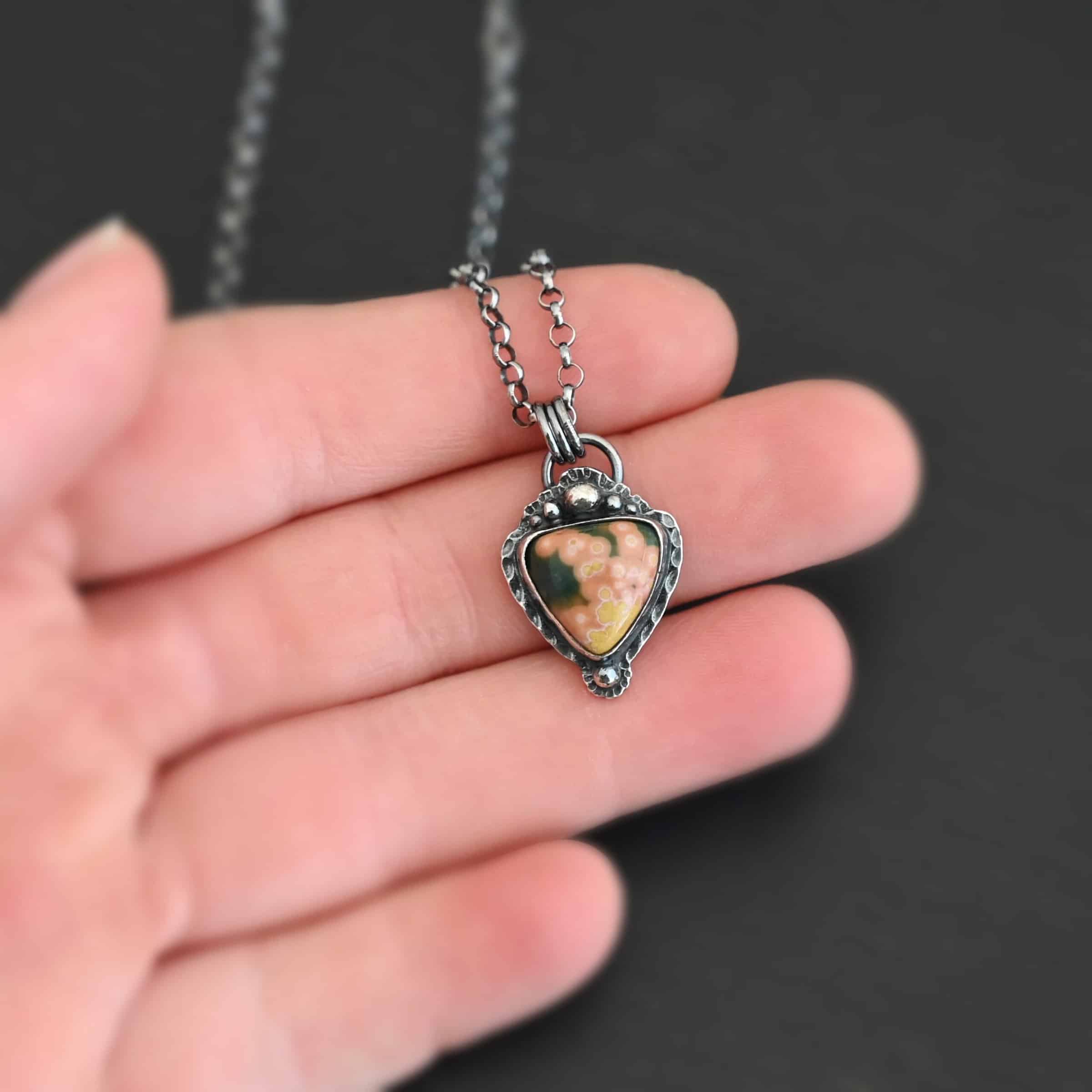 Close up of the "Mesa" Necklace. Made out of sterling silver and ocean jasper. The jasper is a triangle shape, point facing downwards and is primarily orange and yellow with dark green scattered around the top of it. There are five silver dots on the top of the pendant which is flat since the top point of the triangle is facing downwards. There is a silver dot on the bottom of the tip of the necklace. There is a loop on the back of the pendant in which jump rings are attached to. There is a chain attached to the jump rings. Necklace is laying against a dark background.