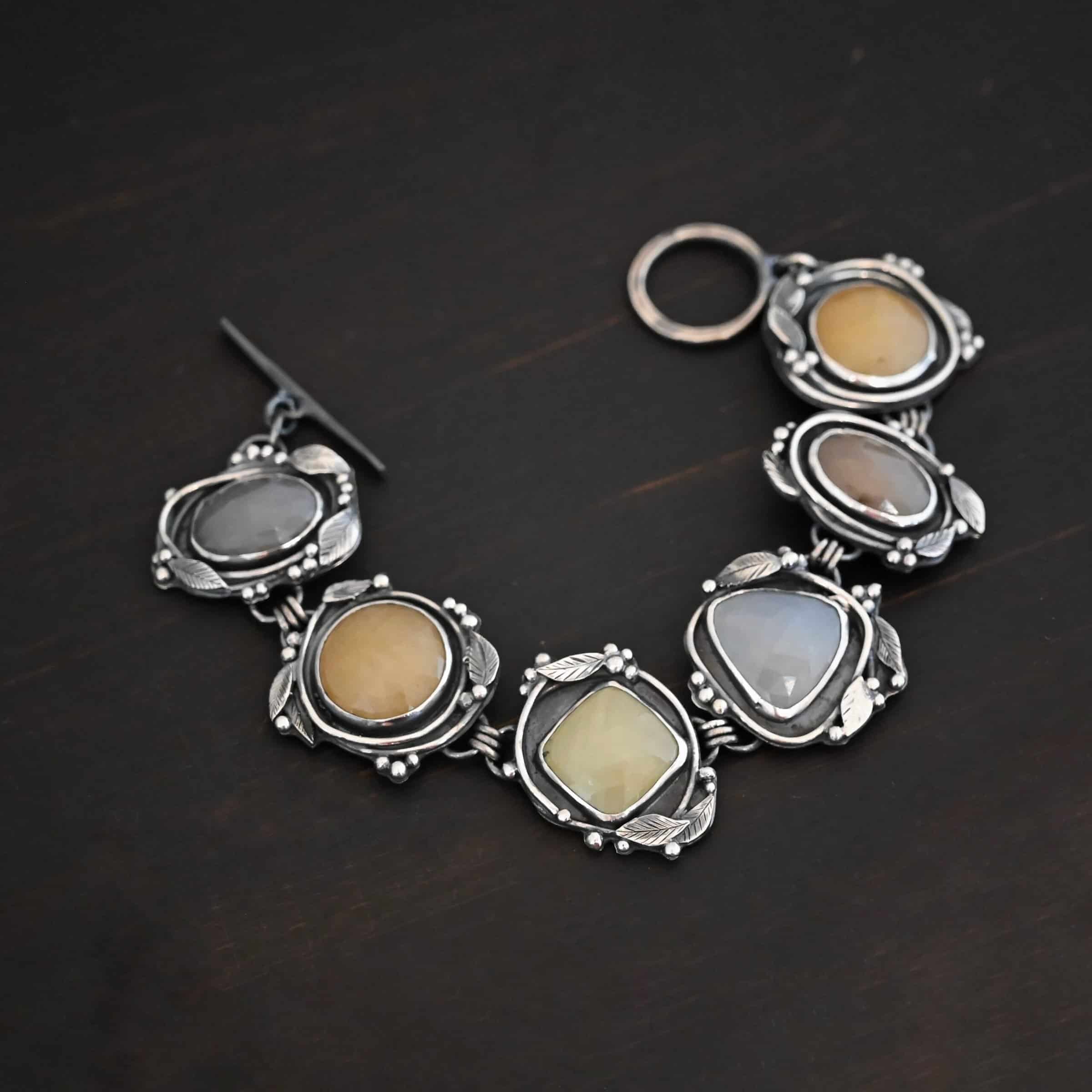 Stone Garden Bracelet Close up of the "Stone Cottage" Bracelet. Made out of sterling silver and sapphires. Each sapphire is a different shape and color. They are either a light gray/blue color, yellow, or brown. Each stone is set in two layers of sterling silver that overlap each other. Each layer of silver has multiple sterling silver leaves and balls around it. Bracelet has a toggle clasp, and is laying against a dark background.