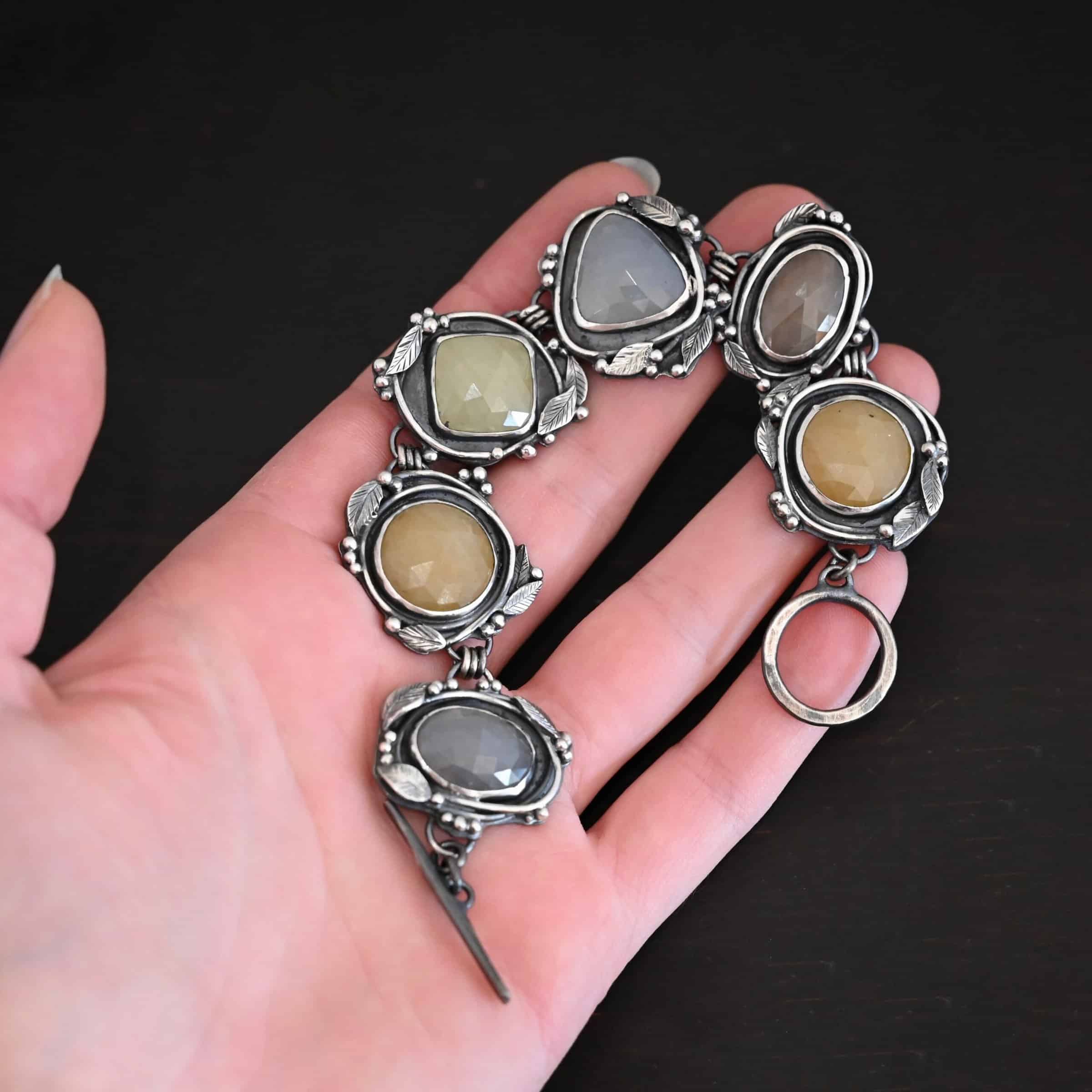 Stone Cottage Bracelet Close up of the "Stone Cottage" Bracelet. Made out of sterling silver and sapphires. Each sapphire is a different shape and color. They are either a light gray/blue color, yellow, or brown. Each stone is set in two layers of sterling silver that overlap each other. Each layer of silver has multiple sterling silver leaves and balls around it. Bracelet has a toggle clasp, and is laying against the palm of a hand to demonstrate size.
