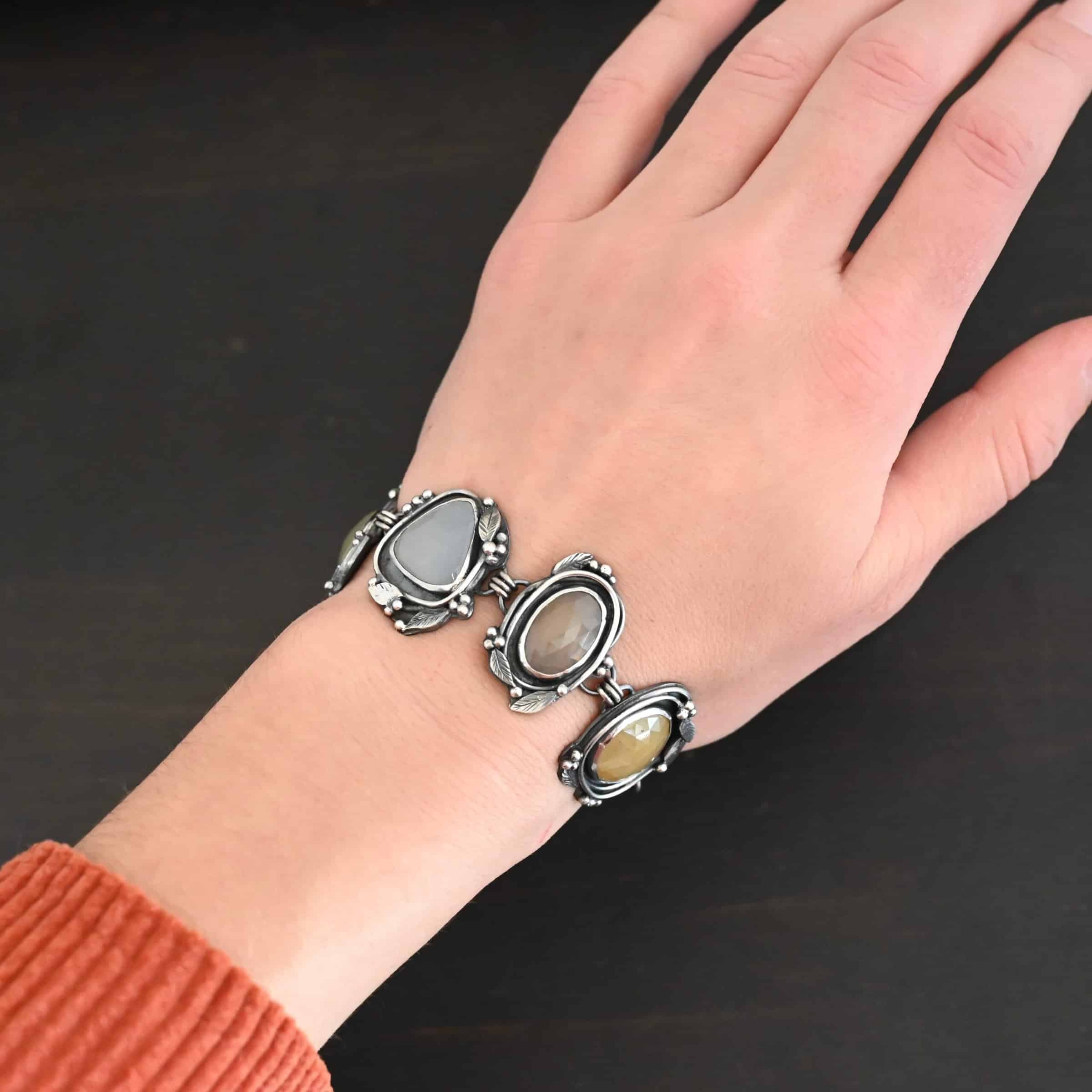 Stone Cottage Bracelet Close up of the "Stone Cottage" Bracelet. Made out of sterling silver and sapphires. Each sapphire is a different shape and color. They are either a light gray/blue color, yellow, or brown. Each stone is set in two layers of sterling silver that overlap each other. Each layer of silver has multiple sterling silver leaves and balls around it. Bracelet has a toggle clasp, and is worn by a model to demonstrate size.