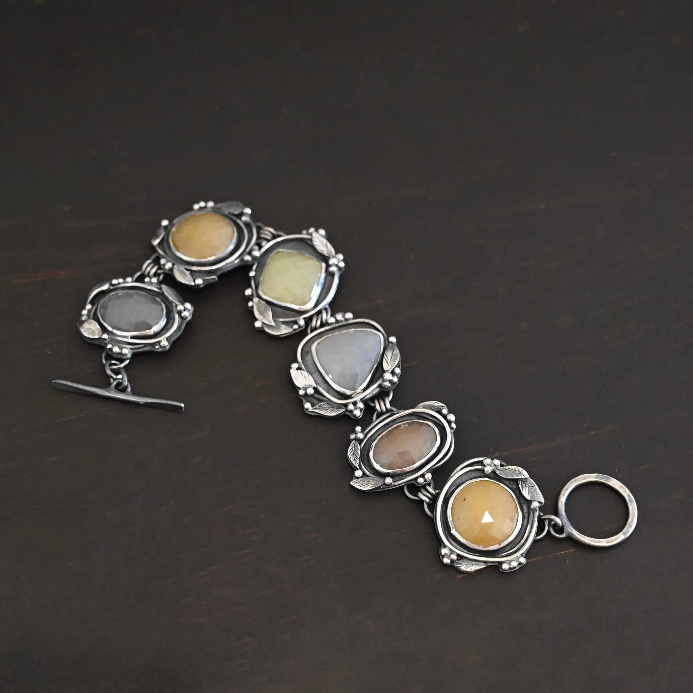 Stone Cottage Bracelet Close up of the "Stone Cottage" Bracelet. Made out of sterling silver and sapphires. Each sapphire is a different shape and color. They are either a light gray/blue color, yellow, or brown. Each stone is set in two layers of sterling silver that overlap each other. Each layer of silver has multiple sterling silver leaves and balls around it. Bracelet has a toggle clasp, and is laying against a dark background.