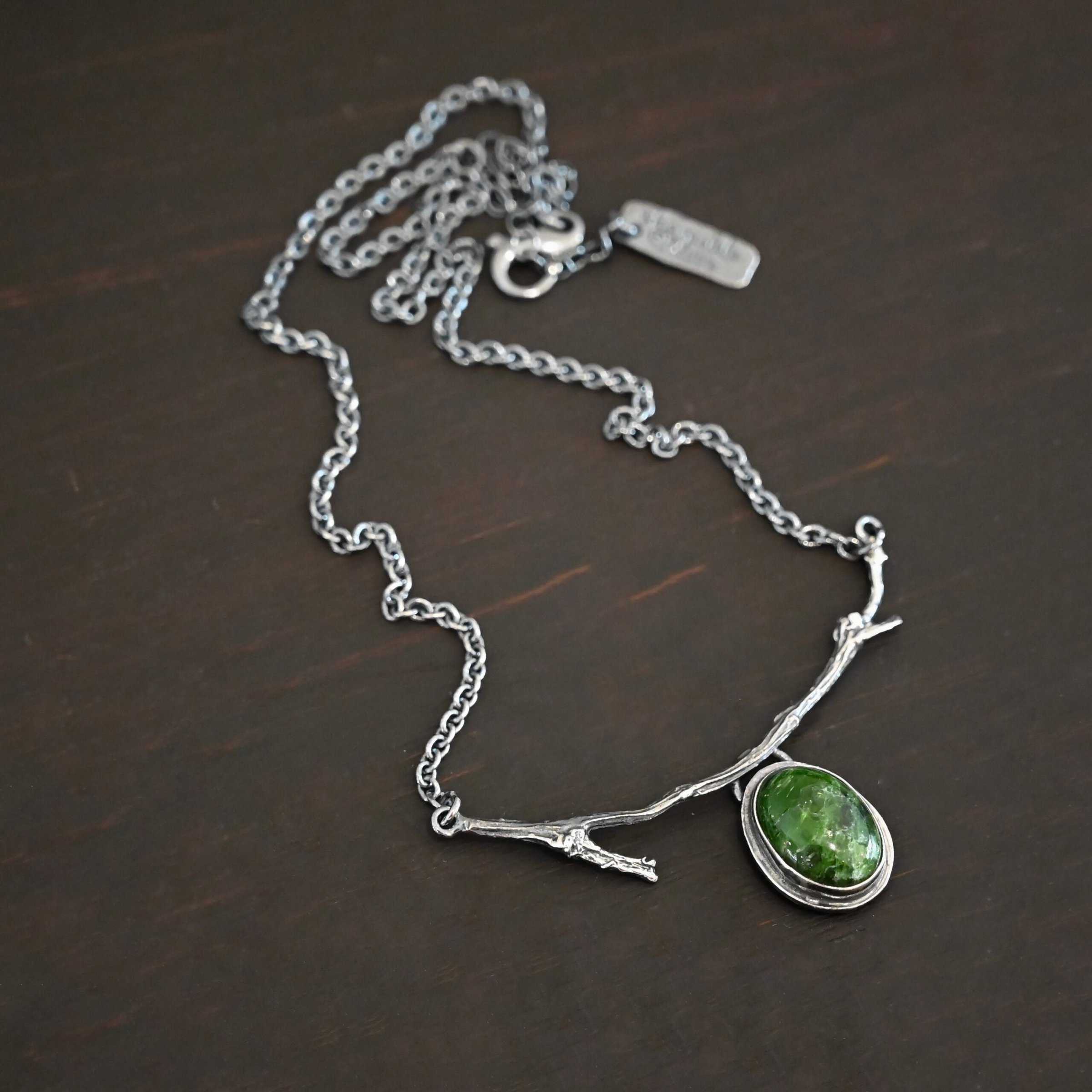 Endless Forest Necklace Close up of the "Endless Forest" Necklace. Made out of sterling silver and chrome diopside. The chrome diopside is an oval shape. It is a dark green color. There is a loop on the back of the pendant that is soldered to a casted "twig" that is curved. Each end of the twig has a loop that a chain is attached to. Each chain leads back to a clasp to connect the necklace. Necklace is shown against a dark background.