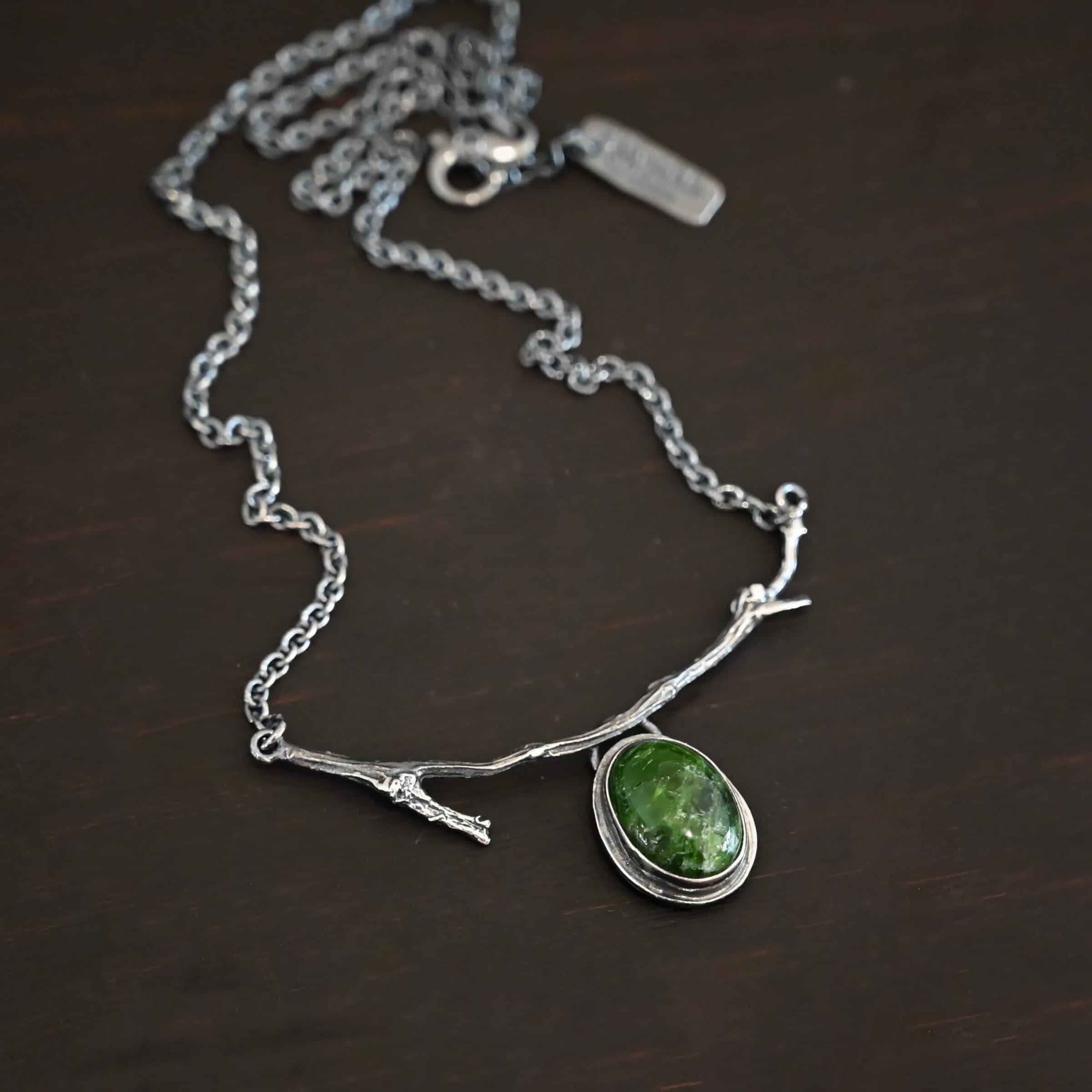 Endless Forest Necklace Close up of the "Endless Forest" Necklace. Made out of sterling silver and chrome diopside. The chrome diopside is an oval shape. It is a dark green color. There is a loop on the back of the pendant that is soldered to a casted "twig" that is curved. Each end of the twig has a loop that a chain is attached to. Each chain leads back to a clasp to connect the necklace. Necklace is shown against a dark background.